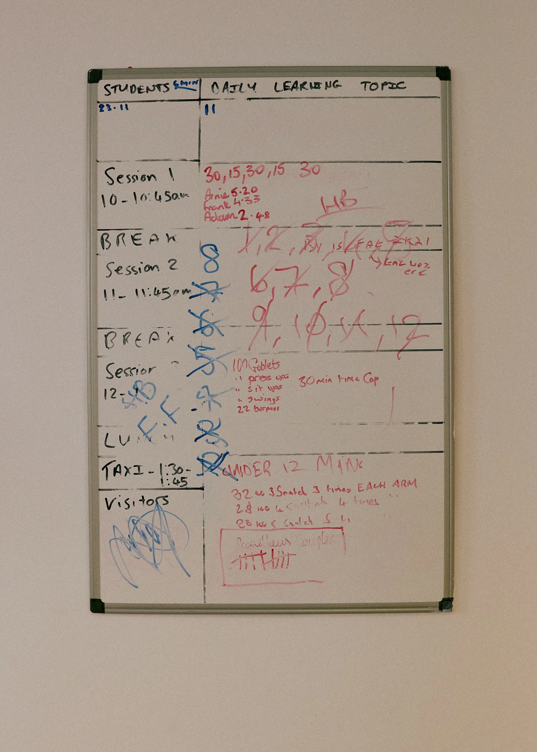 Whiteboard with daily schedule, including sessions, breaks, and notes, written in black, red, and blue marker.