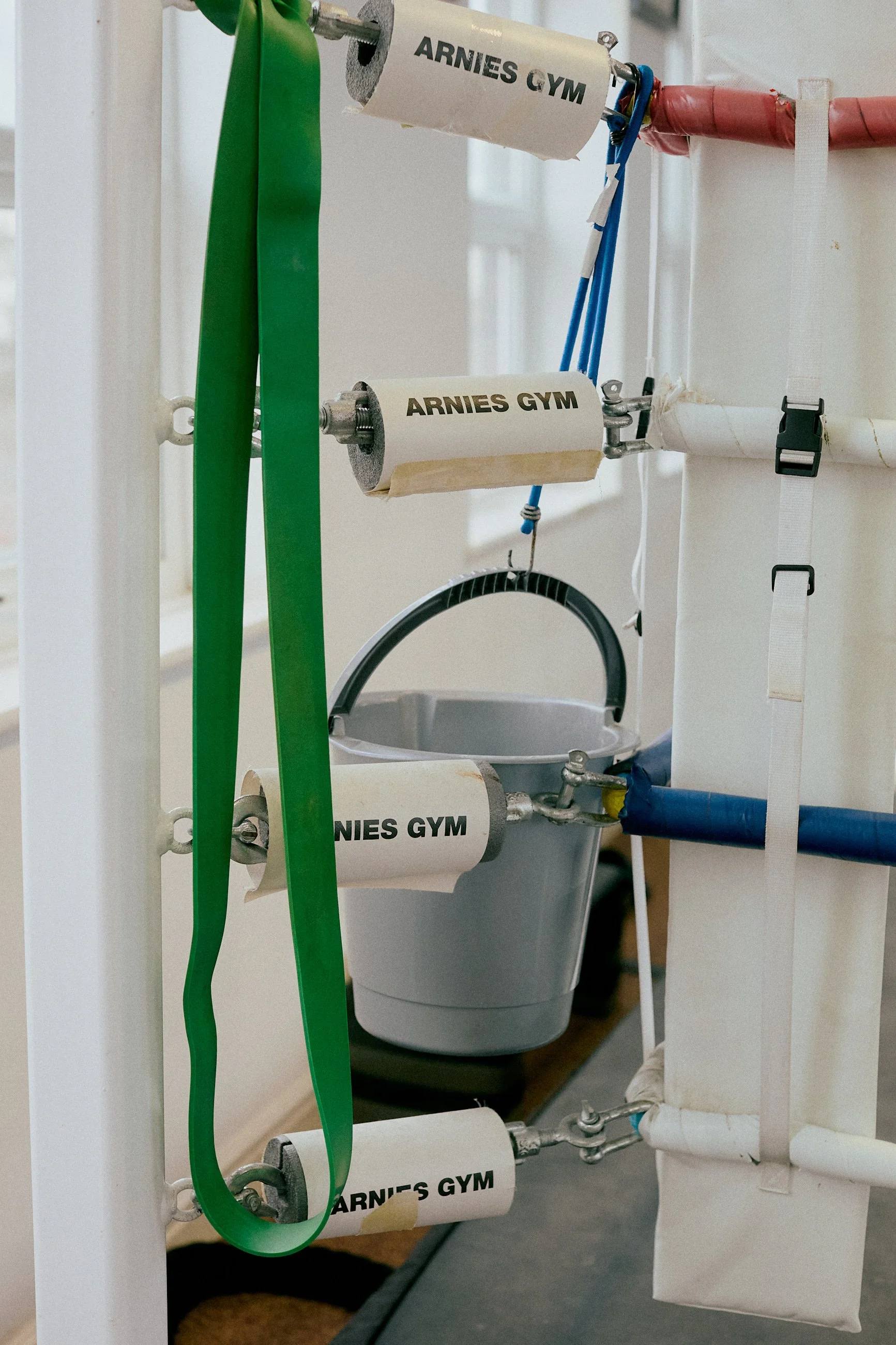 A white gym equipment frame with paper roller labels reading 'ARNIES GYM', green resistance bands, a blue jump rope, and a gray bucket hanging from the frame.