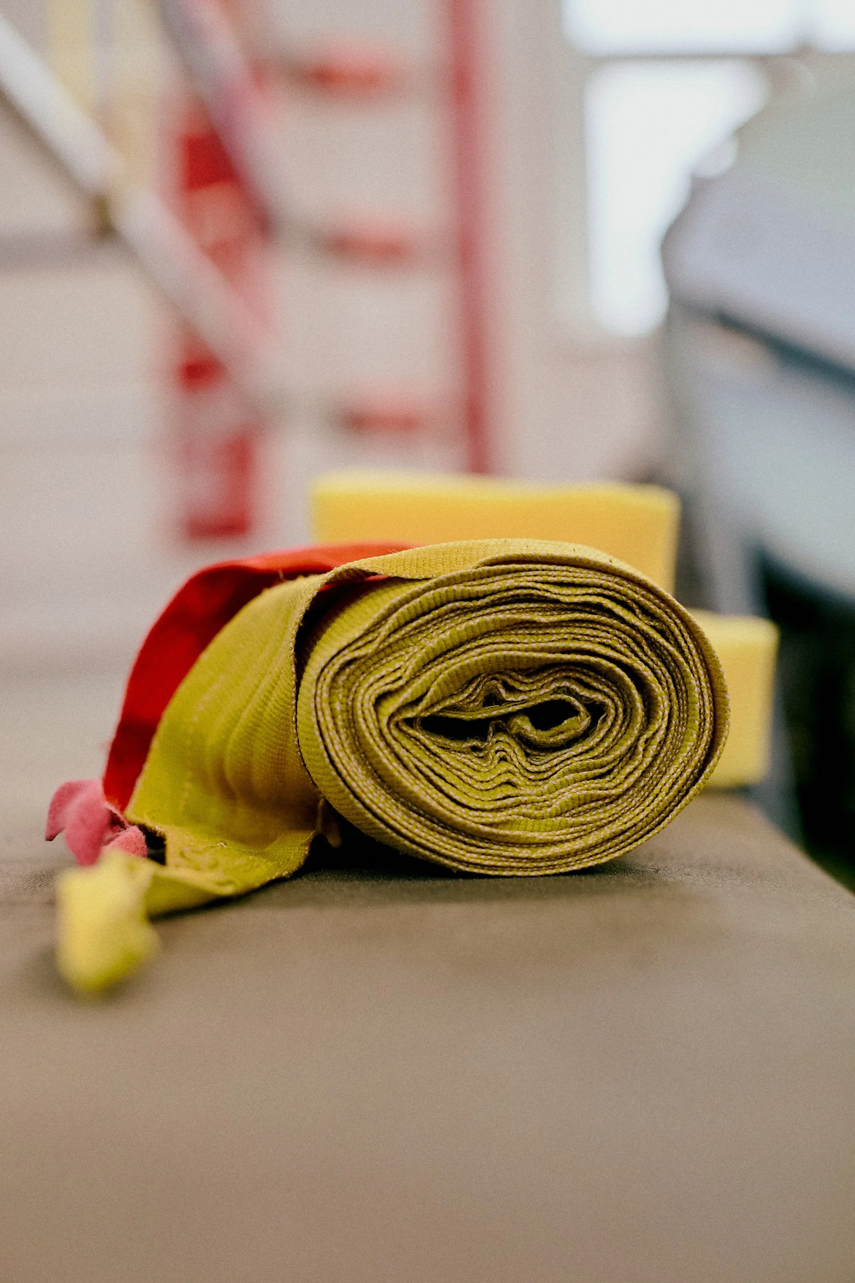 A rolled yellow and red fabric strap resting on a surface with yellow foam blocks and furniture in the background.