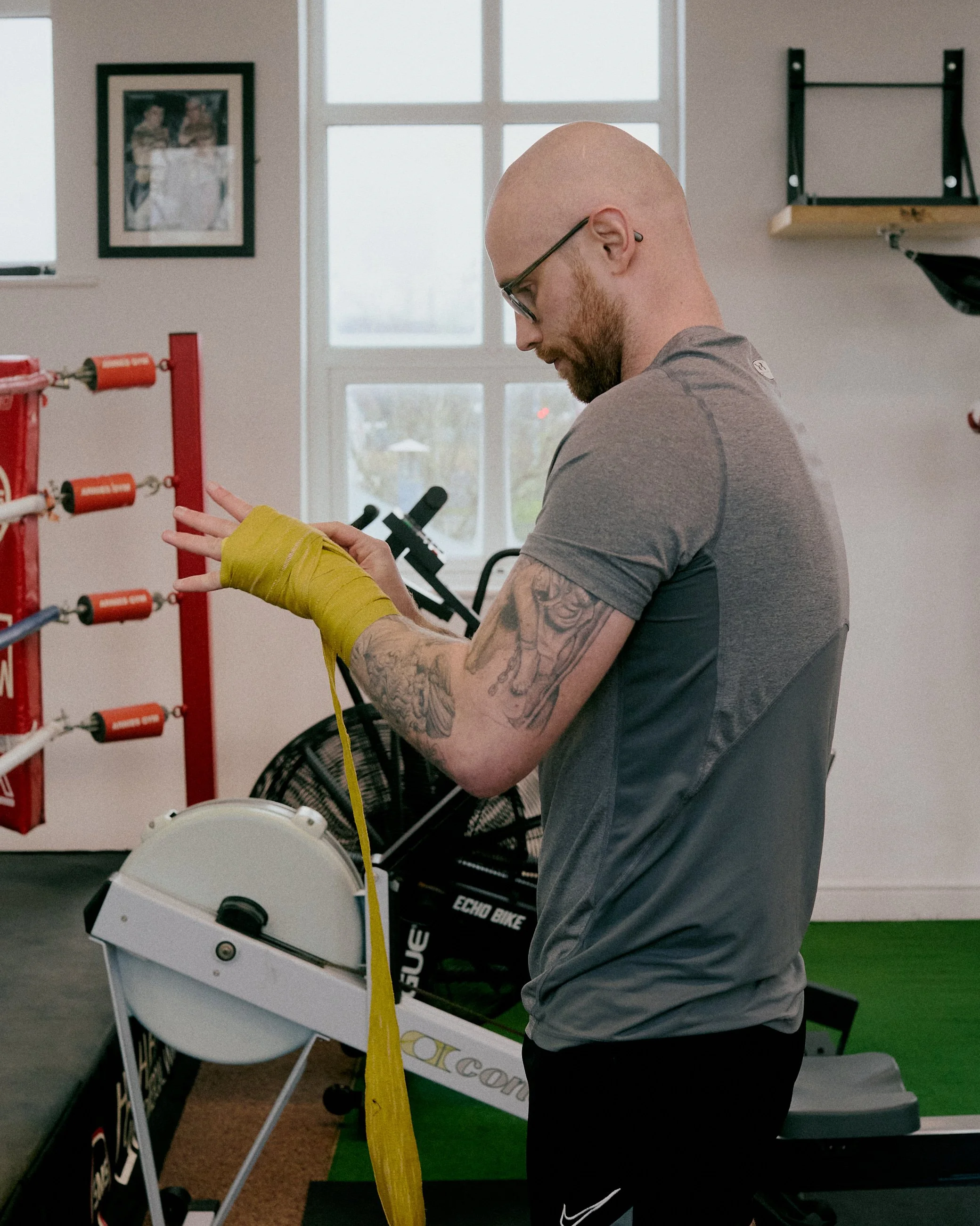 A man with tattoos on his arm and glasses, wrapped in yellow hand wraps, is in a gym preparing for workout or training, standing next to a rowing machine.