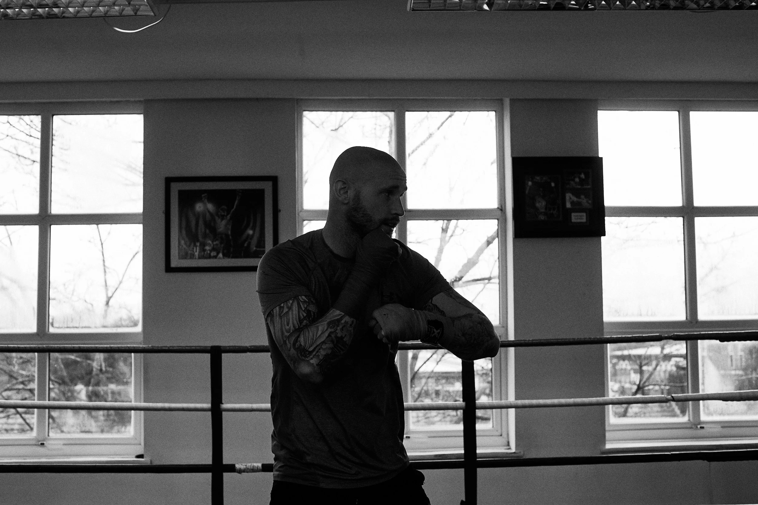 A muscular man with tattoos and a shaved head standing inside a boxing gym near the ring, resting his chin on his hand, looking serious.