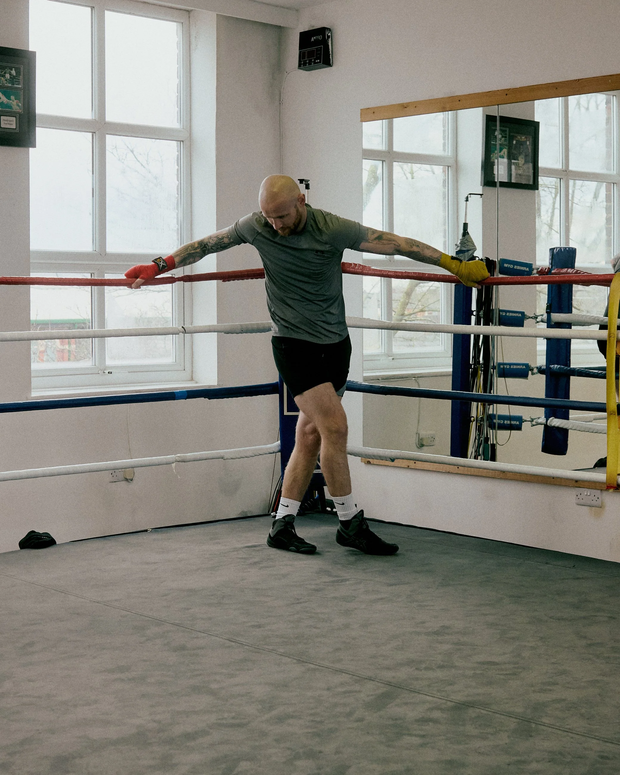 A man in boxing gloves standing in a boxing ring, leaning on the ropes with both arms stretched out, looking down, inside a gym with large windows.