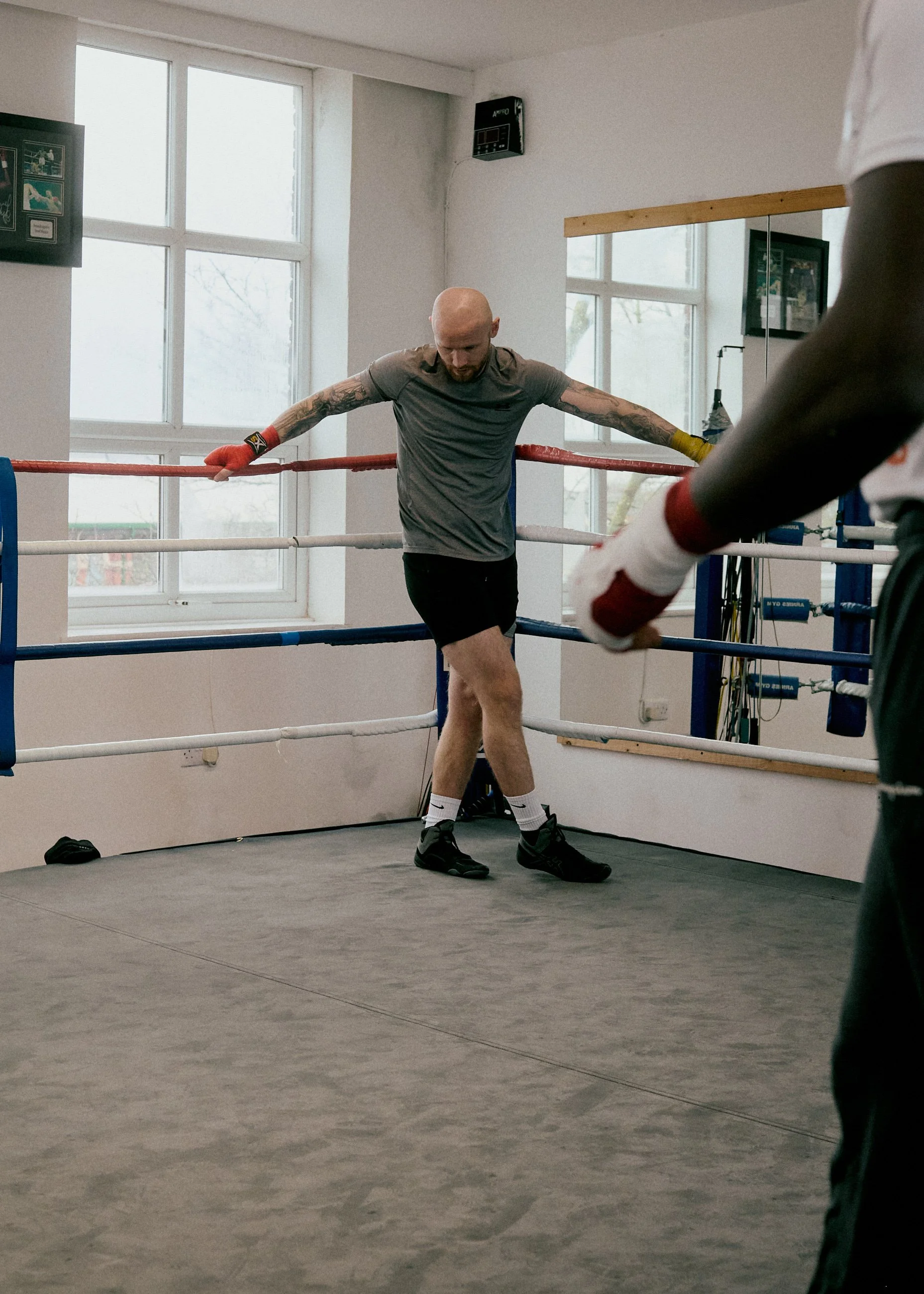 A man with tattoos is in a boxing gym, standing in a boxing ring with his head bowed and arms resting on the ropes, wearing boxing gloves, a gray t-shirt, black shorts, and black shoes.