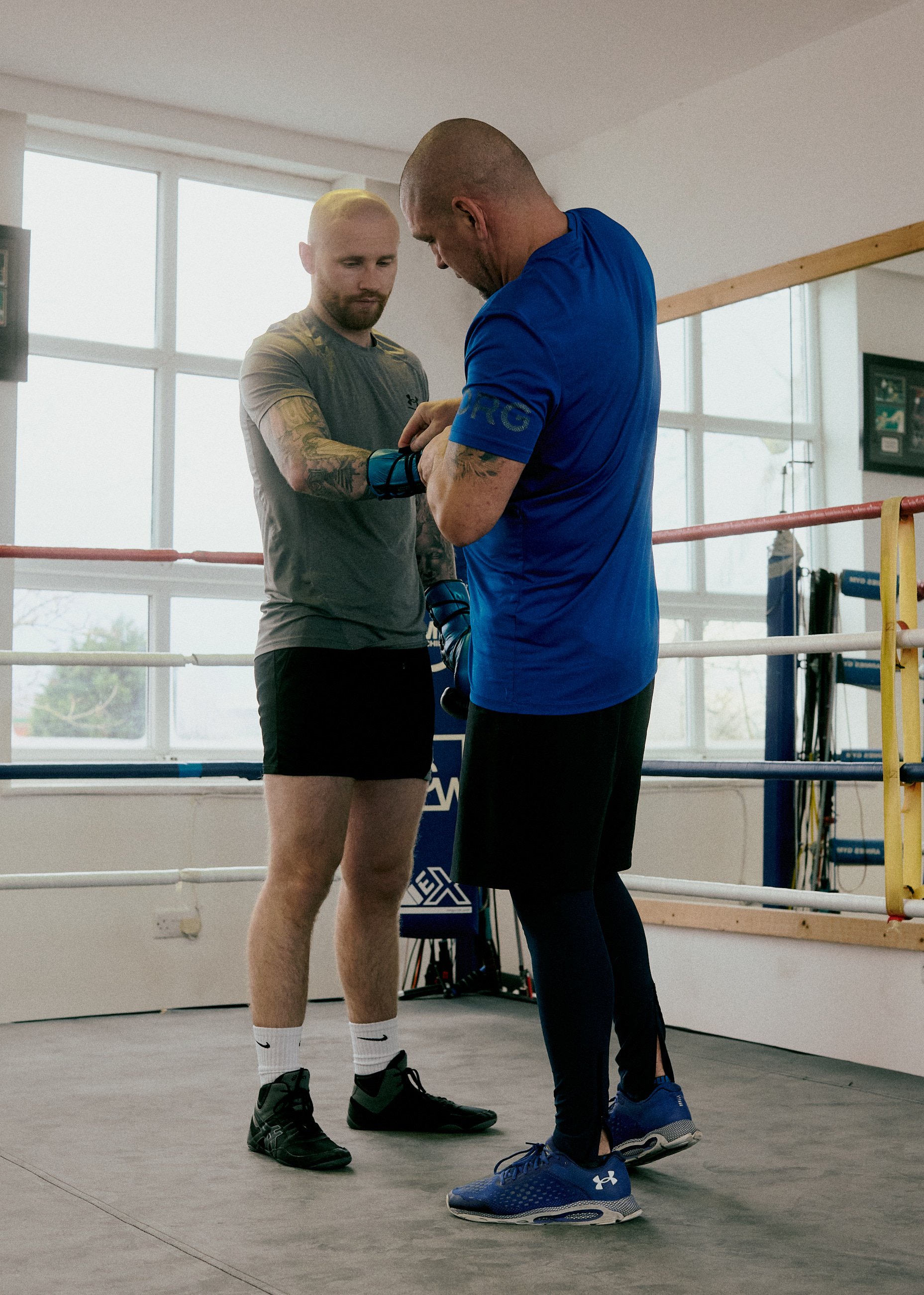 Two men in athletic wear inside a boxing gym. One man is helping the other put on fingerless boxing gloves.