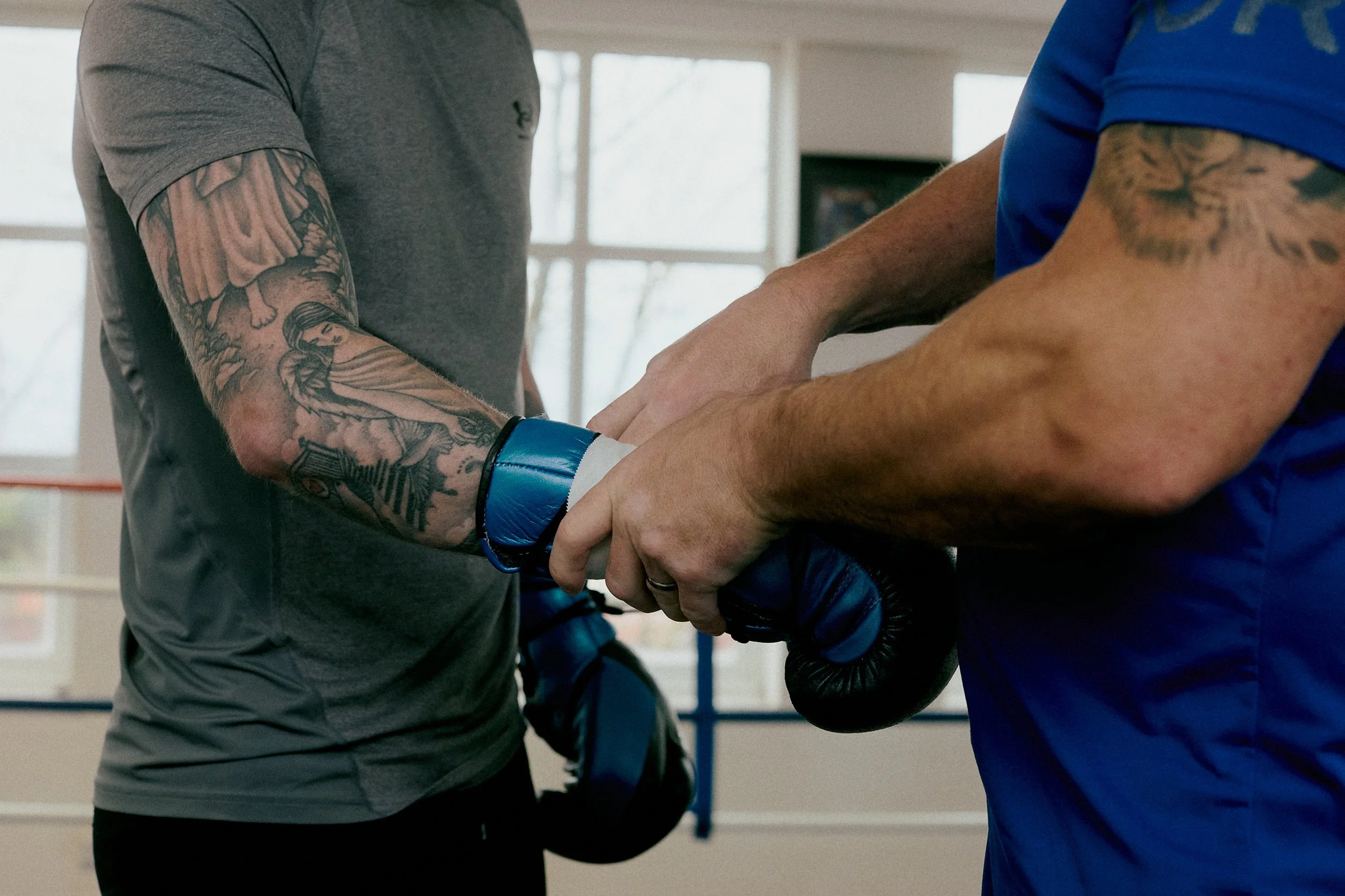 A person in a gray t-shirt with tattoos on their arm is holding a blue boxing glove with another person in a blue t-shirt, also with tattoos, during a boxing training session indoors.