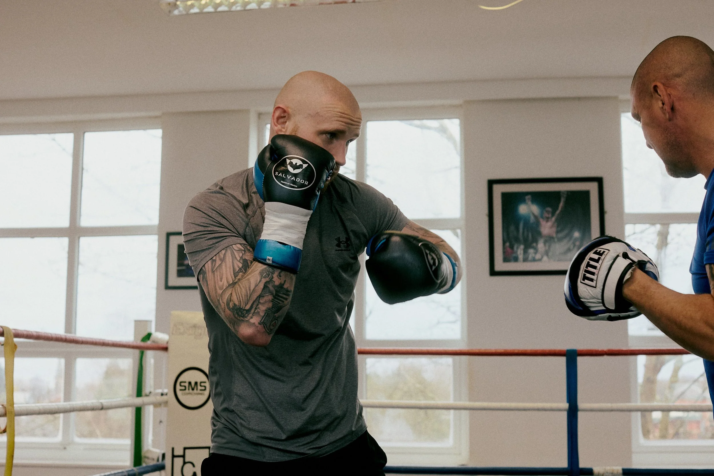 A boxer in a training ring wearing black gloves is practicing punches with a trainer. The boxer has tattoos on his arm and is focusing on the trainer, who is wearing blue gloves.