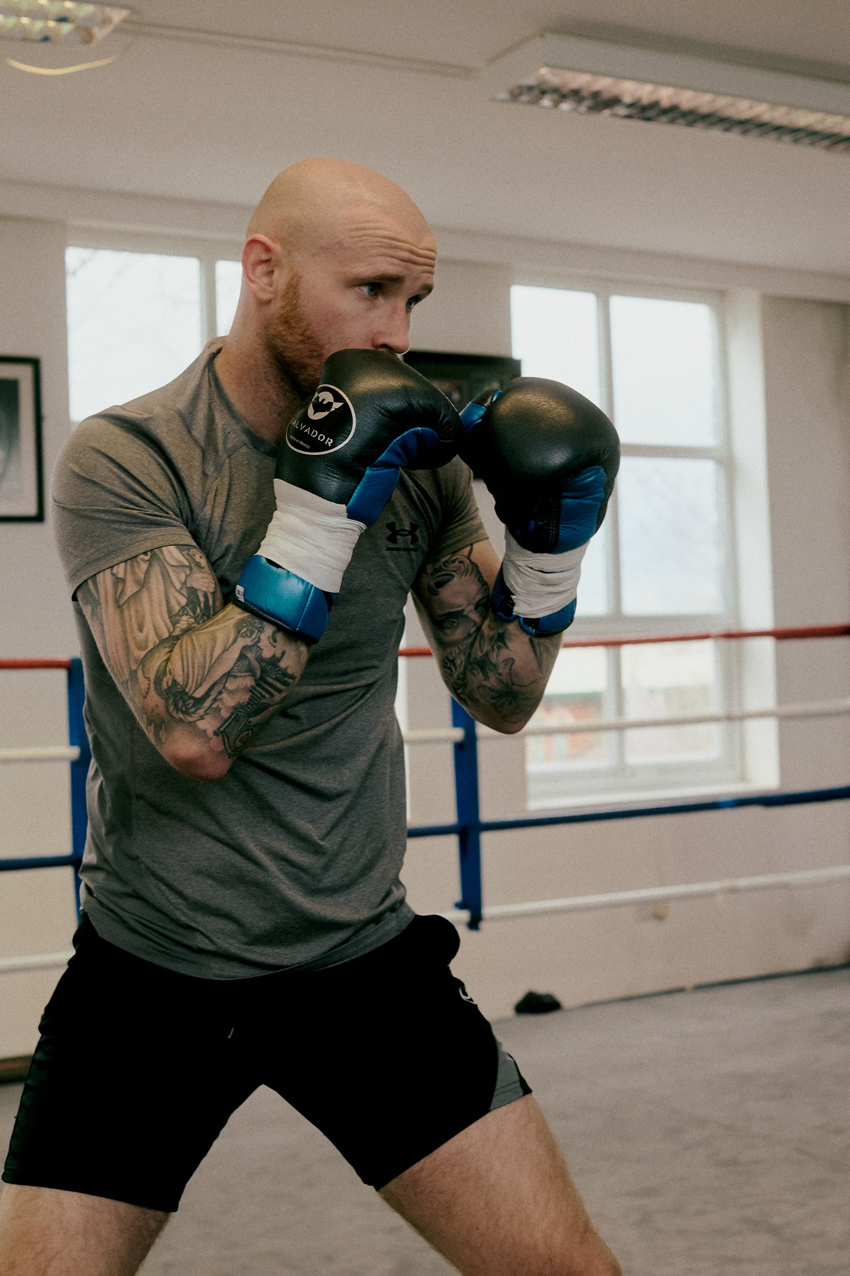 A man with tattoos in a gray athletic shirt and black shorts practicing boxing in a gym, wearing boxing gloves and standing in a ready stance.