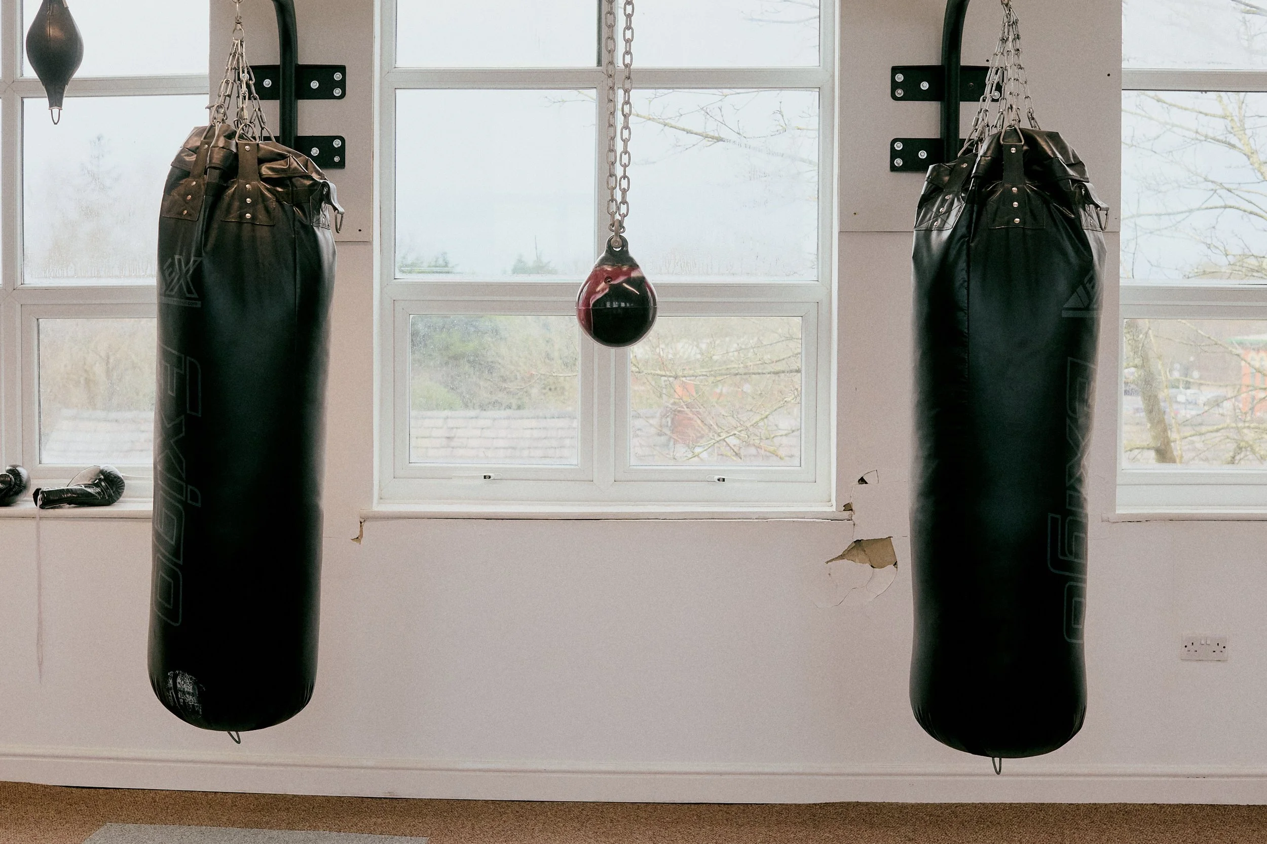 Two black punching bags hanging in a room with a large window in the background, and a red punching ball between them.