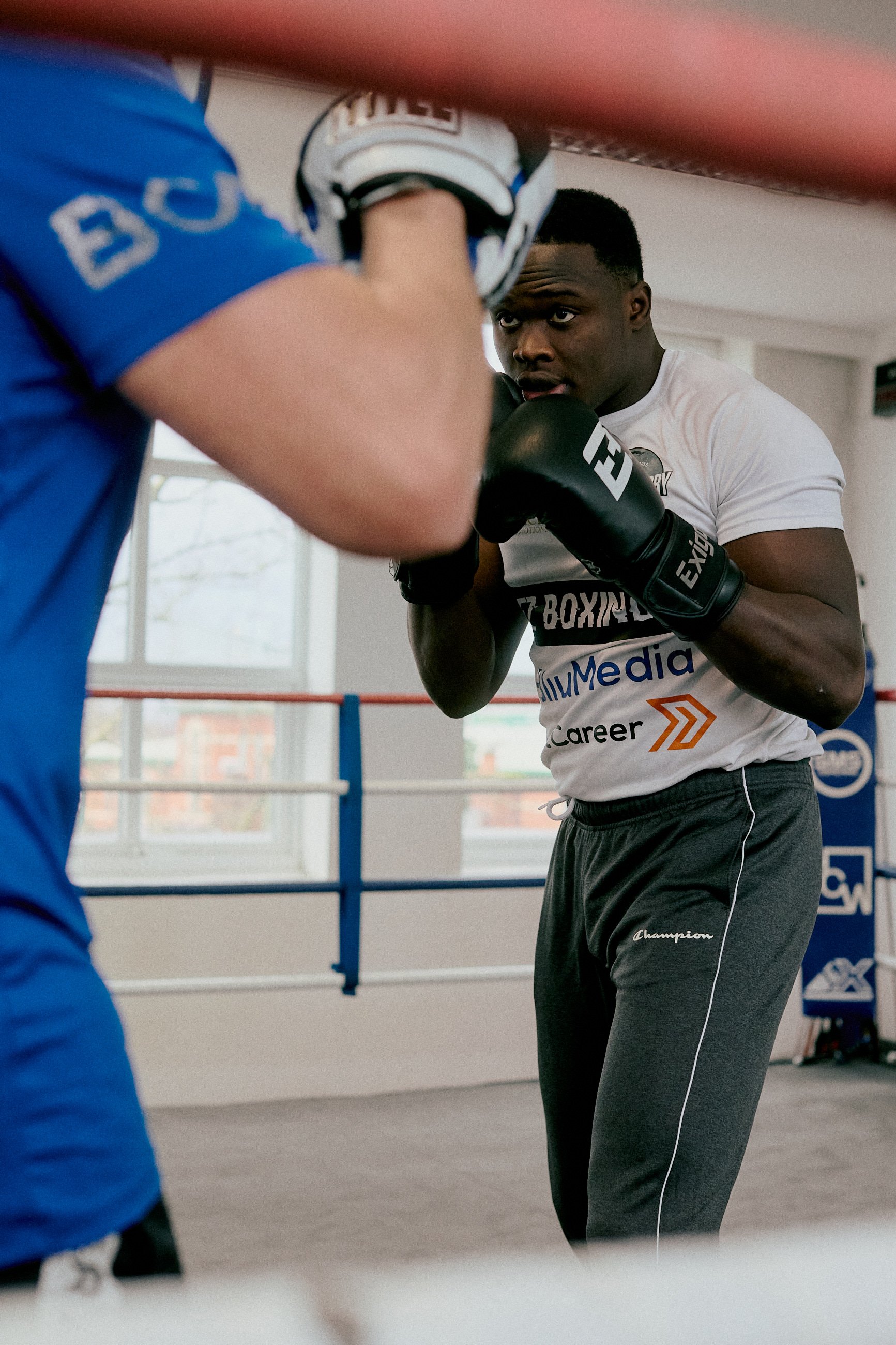 A young boxer in white t-shirt and black gloves sparring with a trainer in a boxing gym.