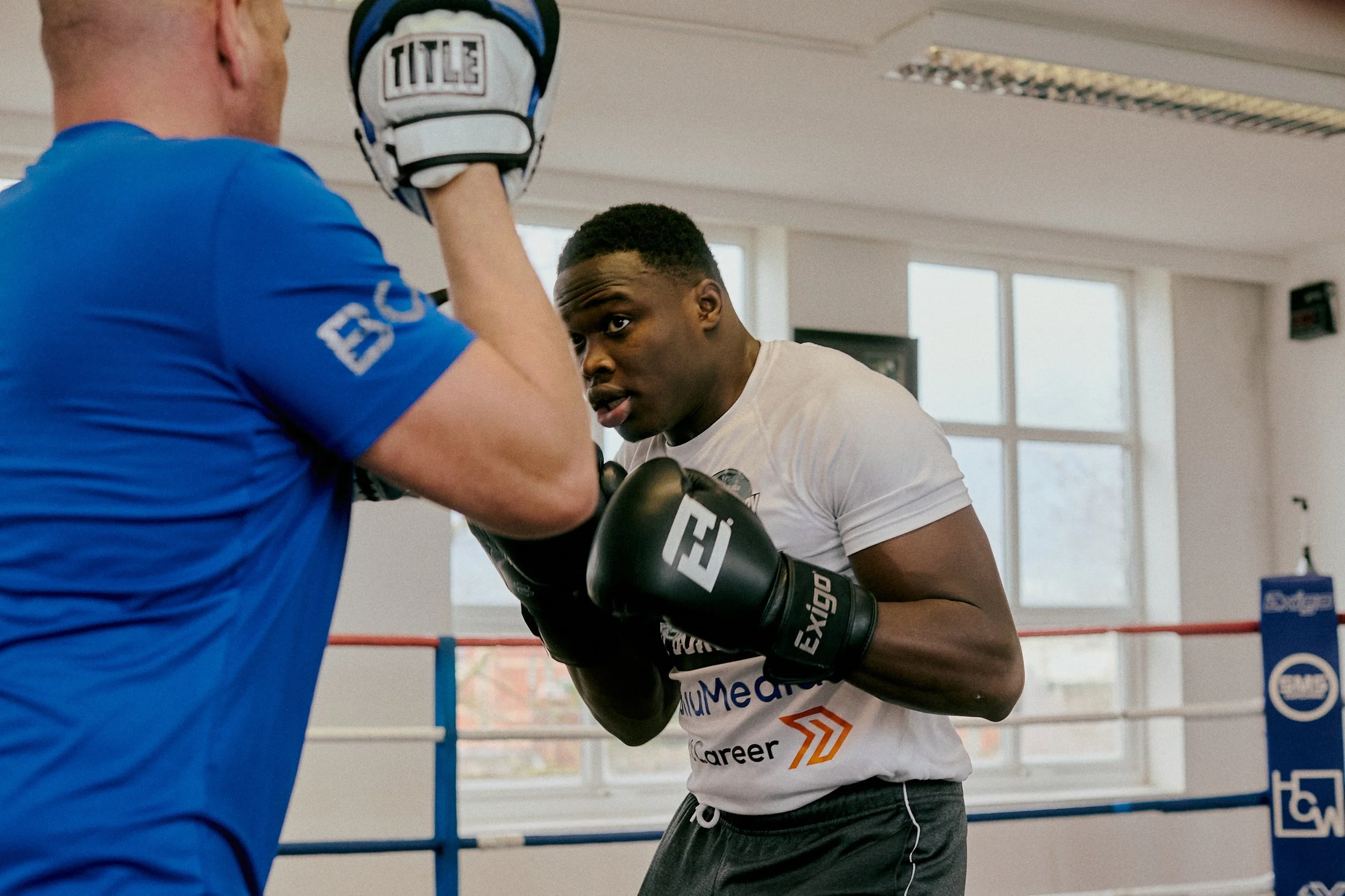 A young male boxer in white t-shirt and black boxing gloves is sparring with a coach in a boxing gym, with red, white, and blue boxing ring ropes in the background.