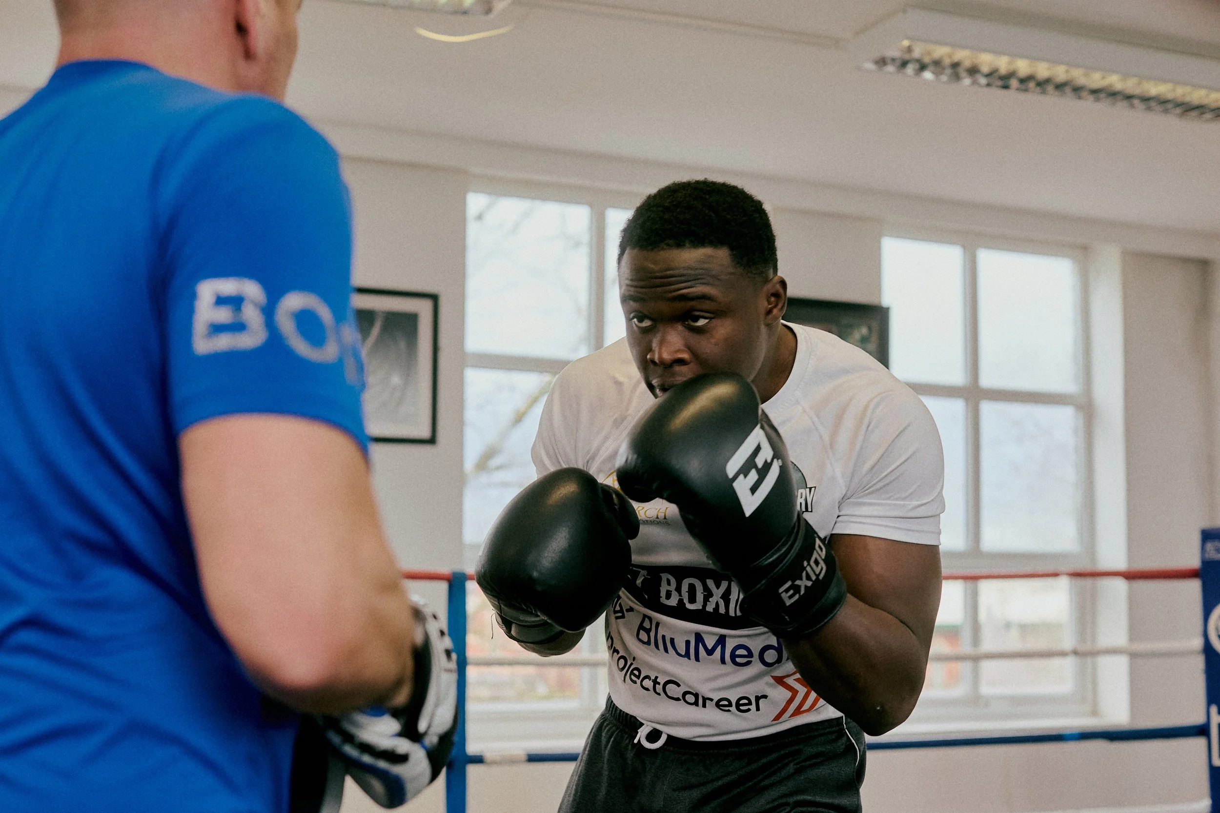 A man wearing boxing gloves practicing boxing in a gym with another person in front of him, wearing a blue shirt.