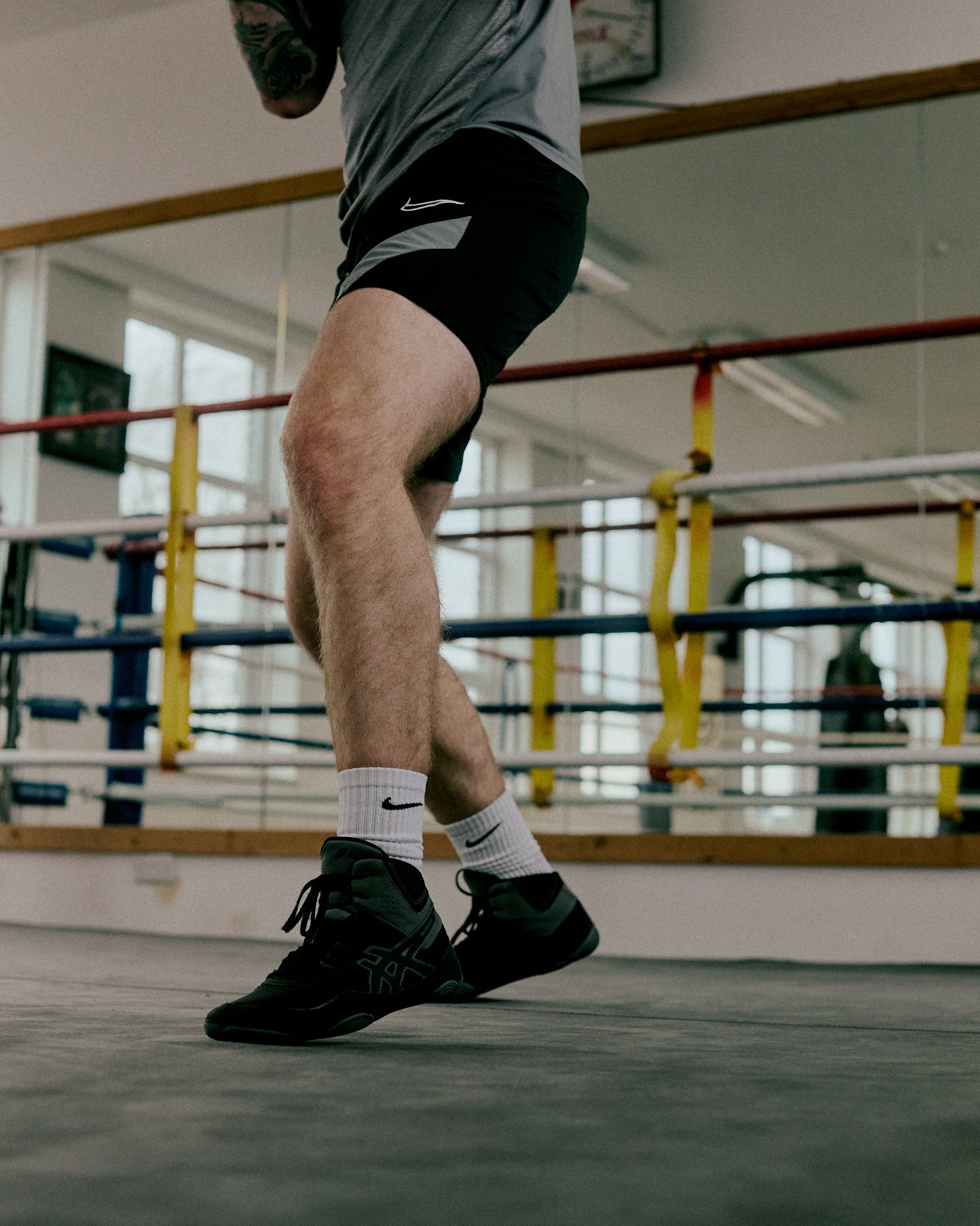 Close-up of a male athlete's legs in black athletic shorts, wearing black running shoes and white Nike socks, practicing jump rope in a gym.