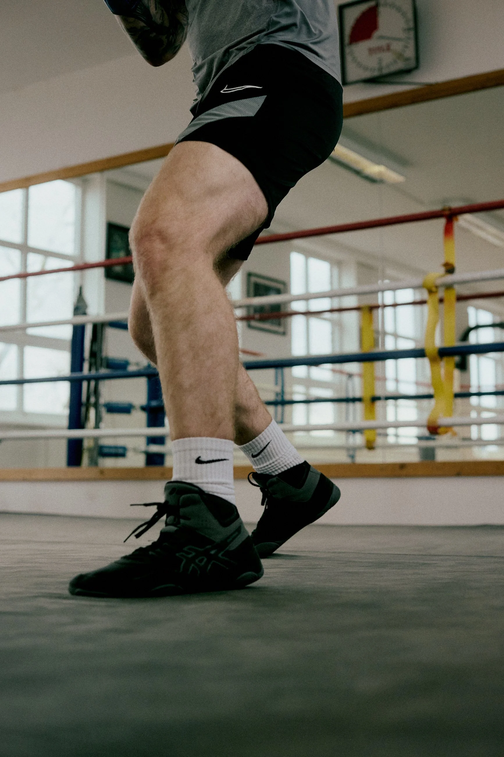 A person in athletic clothing and black sneakers practicing boxing or kickboxing in a gym with boxing ring ropes and large windows.