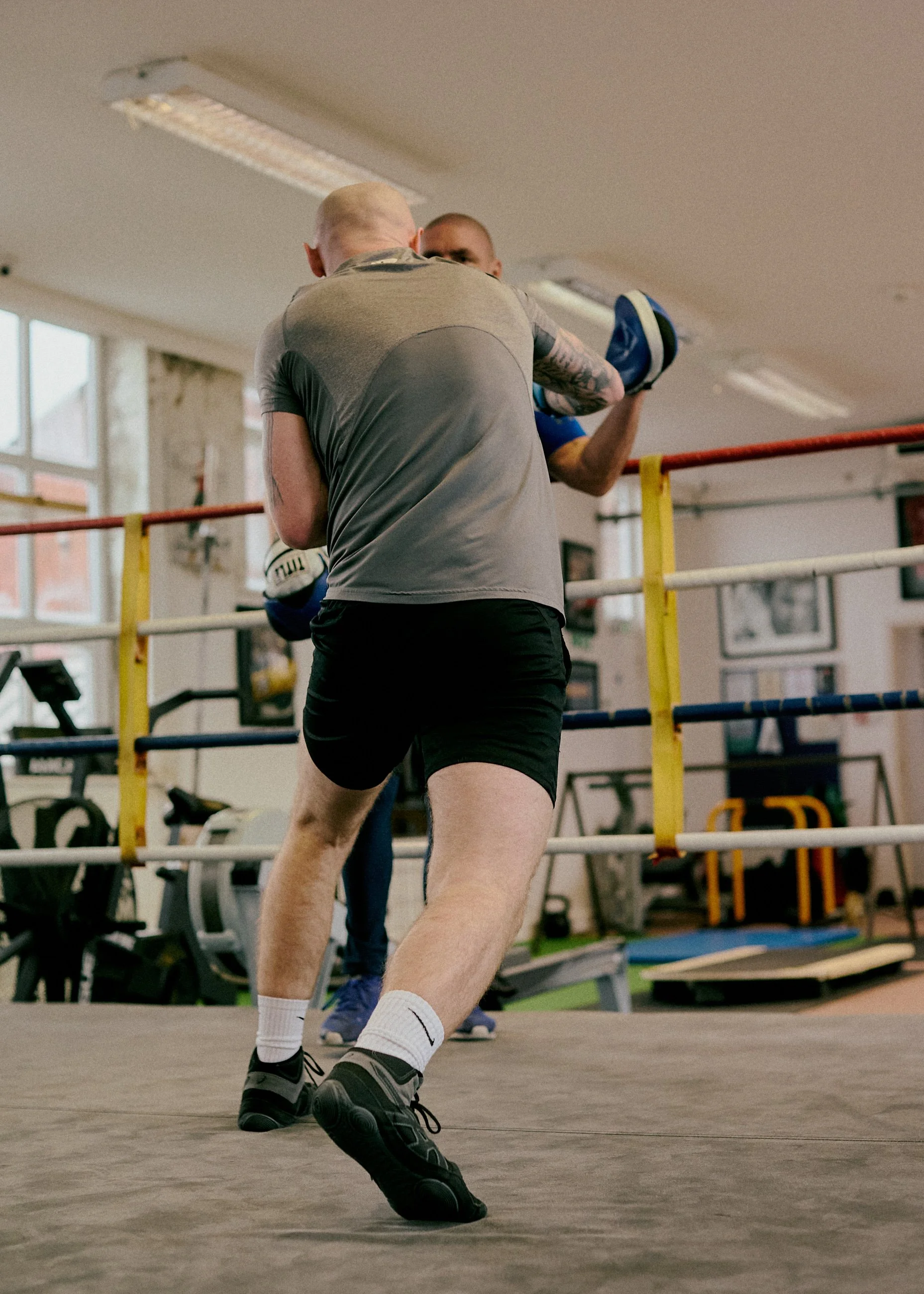 Two men training in a boxing gym, with one man in gray shirt and black shorts practicing punches with a punching bag, and the other man in blue shirt holding the bag.