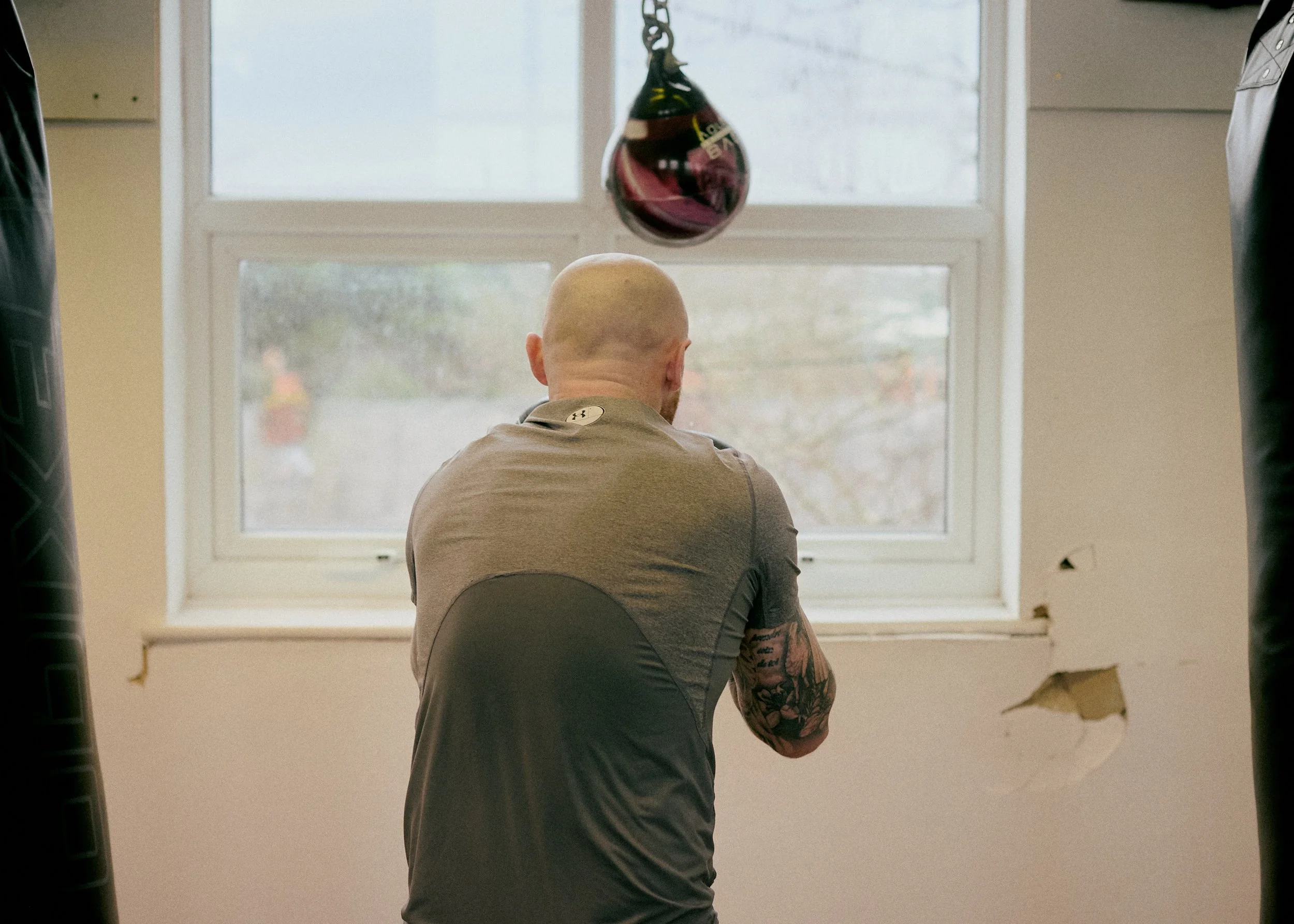 A man with a shaved head, wearing a gray athletic shirt with tattooed arms, sits in a room with walls that have peeling paint and holes. He faces a window where a punching bag hangs in the foreground.