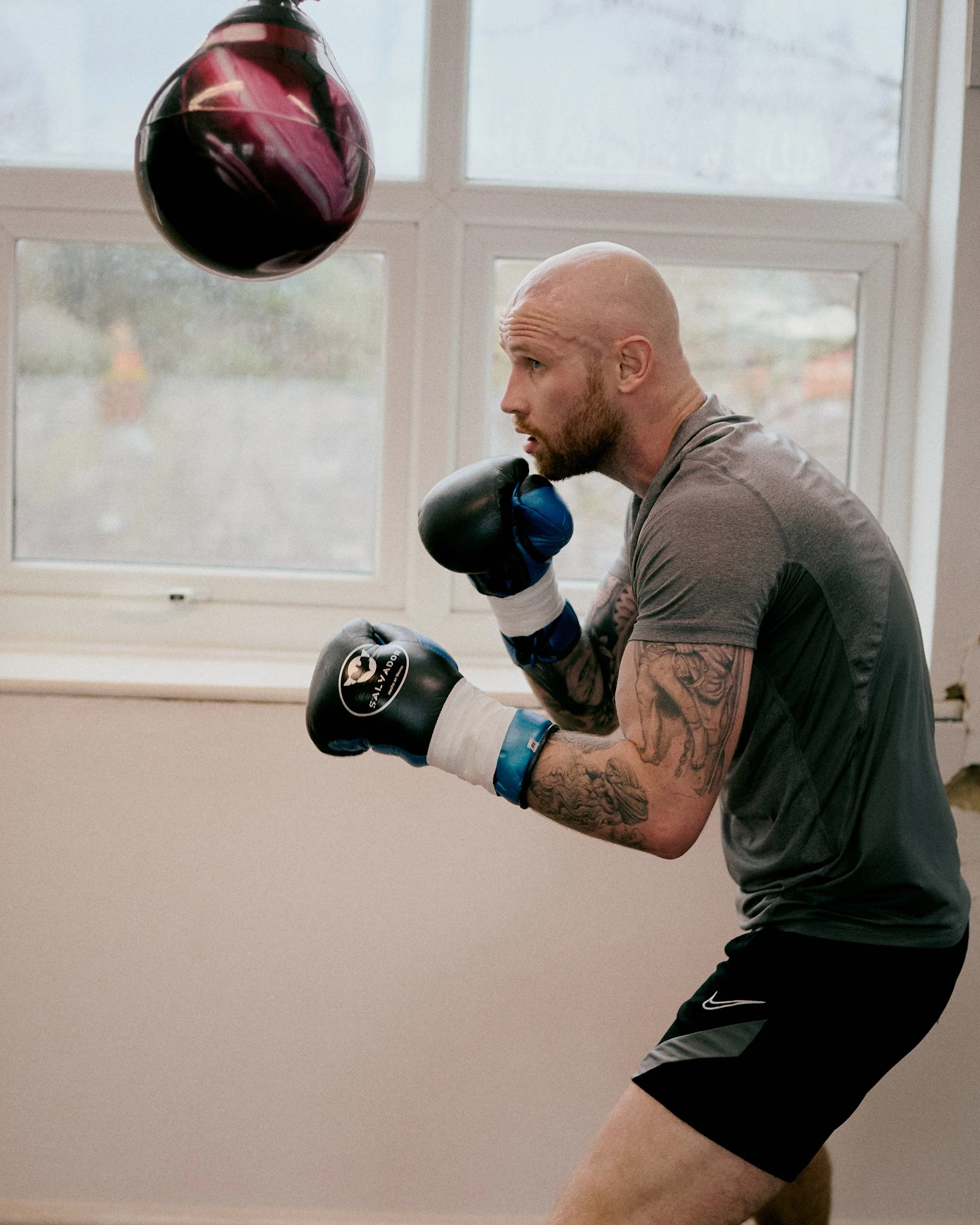 A man with tattoos wearing boxing gloves in a boxing stance indoors, with a focus mitts swinging toward him, in front of a window.