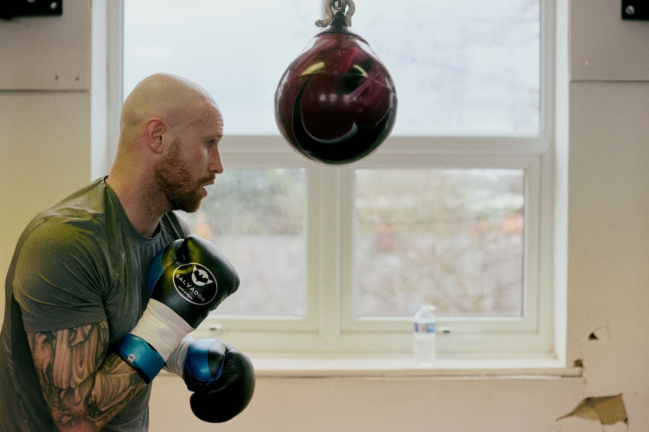 A man with a shaved head and tattoos on his right arm wearing boxing gloves in a gym, practicing boxing or training with a hanging punching bag in front of a window.