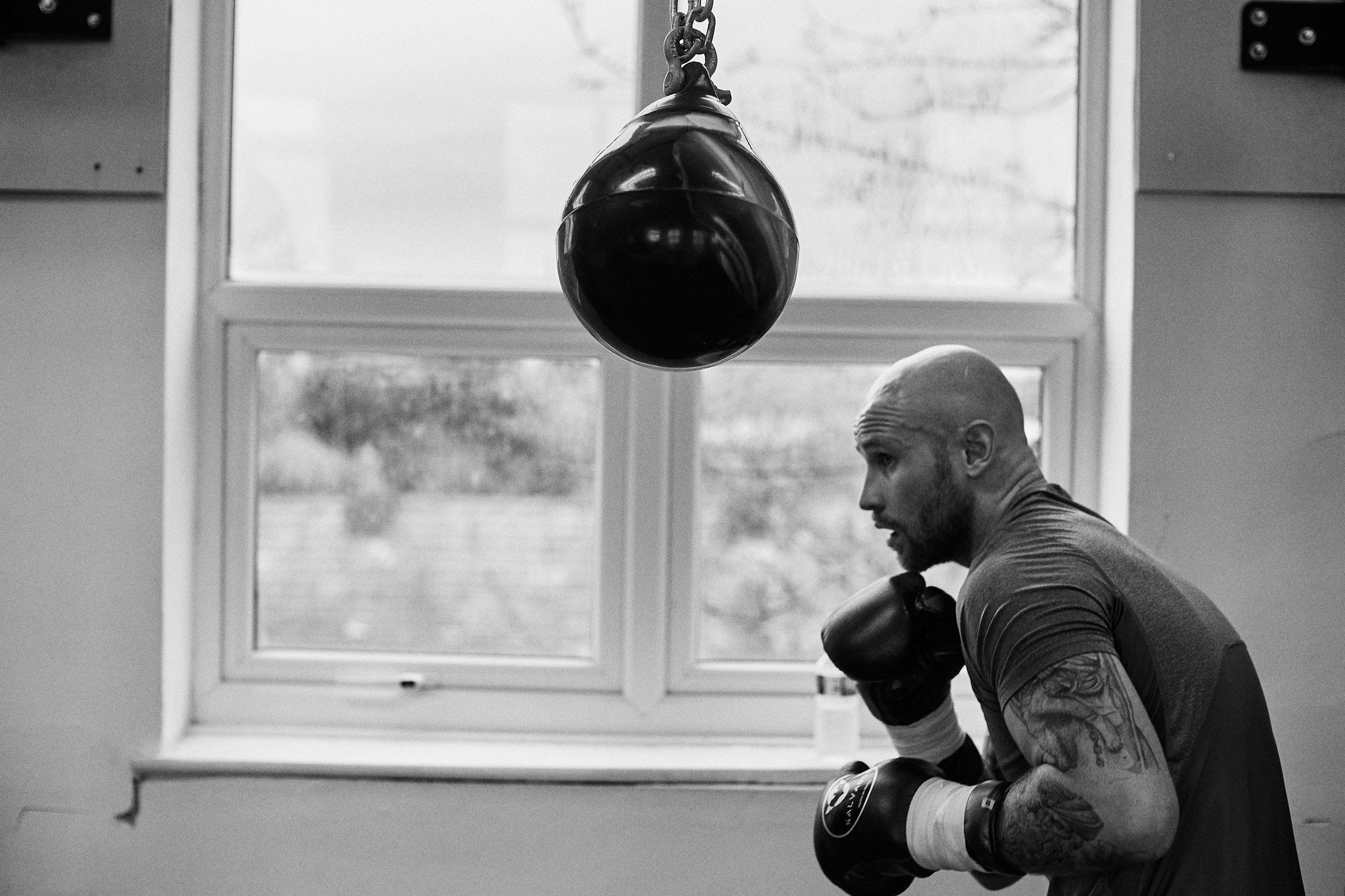 A man practicing boxing in a gym, facing a hanging punching bag, with a window in the background.