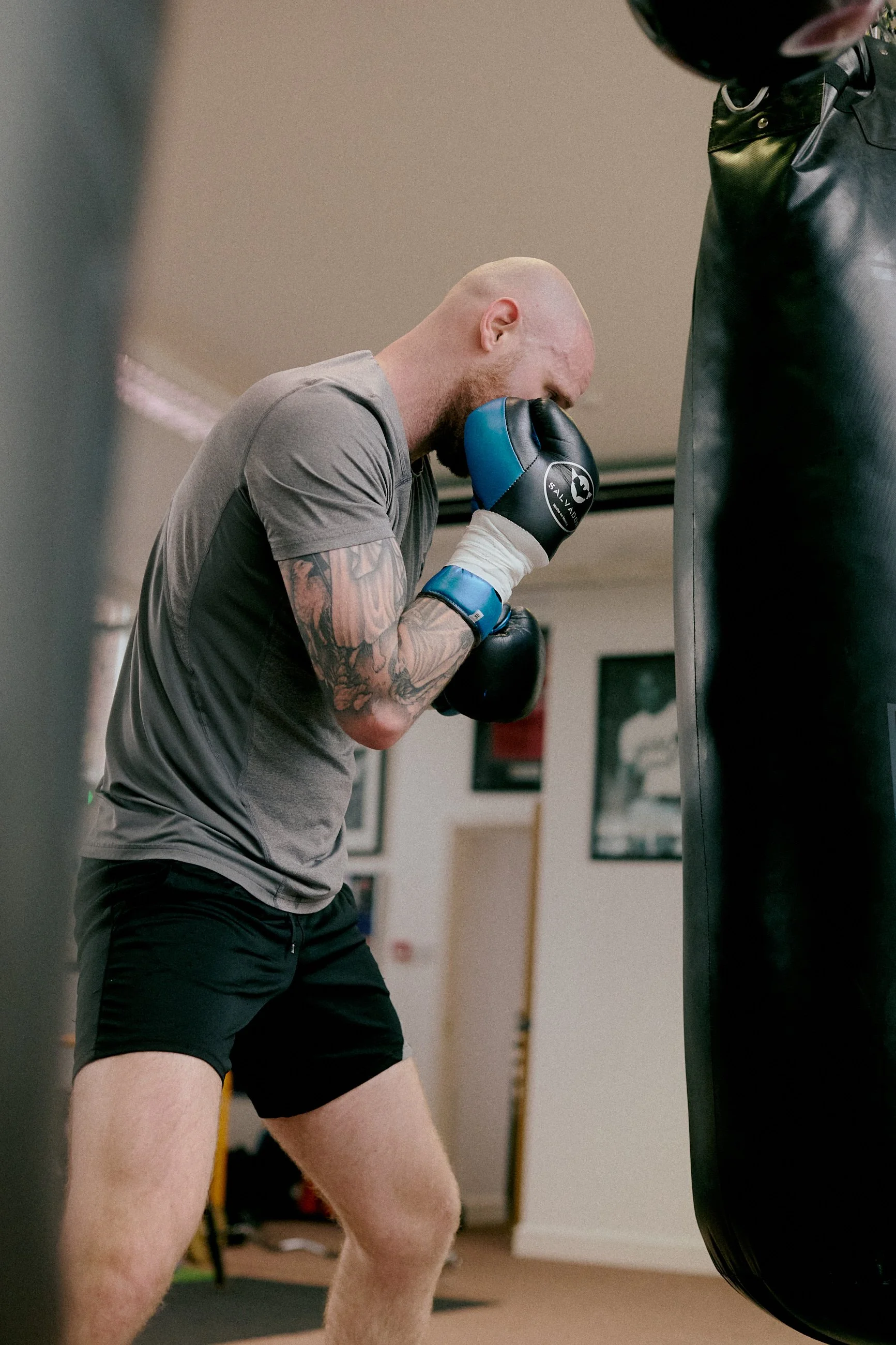 A bald man with tattoos on his left arm, wearing a gray T-shirt and black shorts, practicing boxing on a punching bag while wearing boxing gloves.