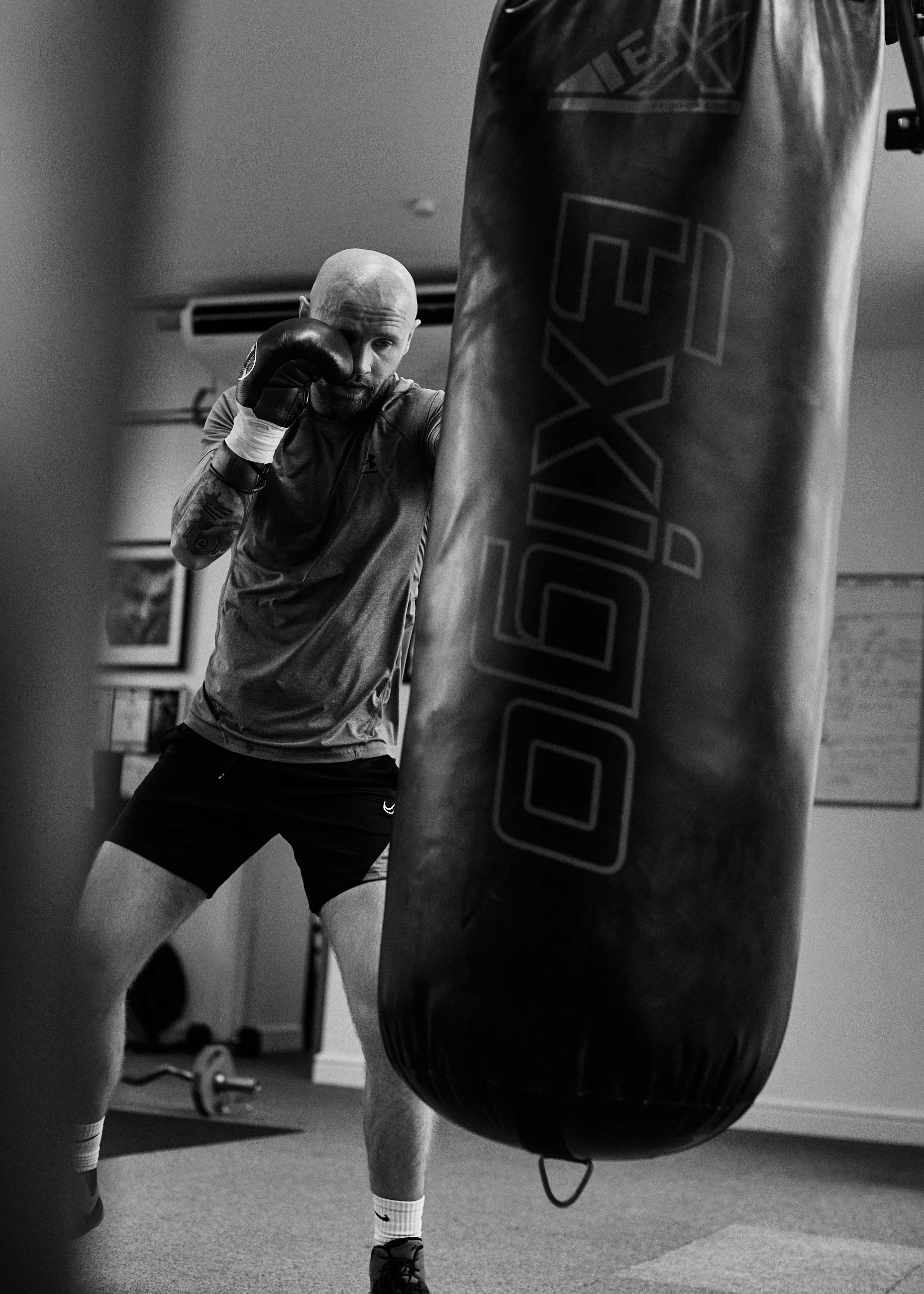 A man with a bald head and tattoos on his arm is practicing boxing with a punching bag in a gym, wearing gloves and athletic clothes.