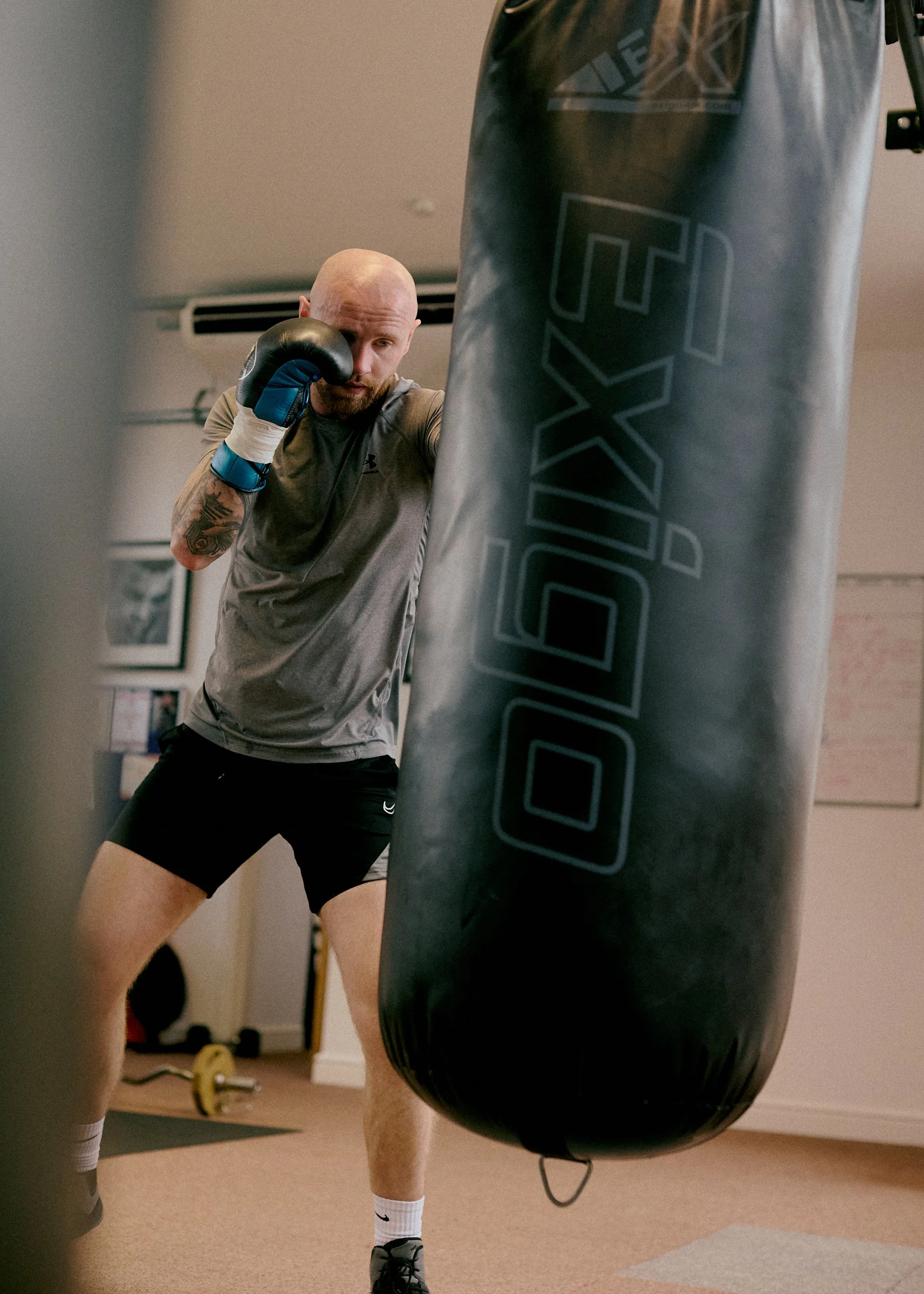 A man with a bald head and tattoos on his arm wearing gray athletic shirt, black shorts, and boxing gloves practicing punches on a black punching bag in a gym.