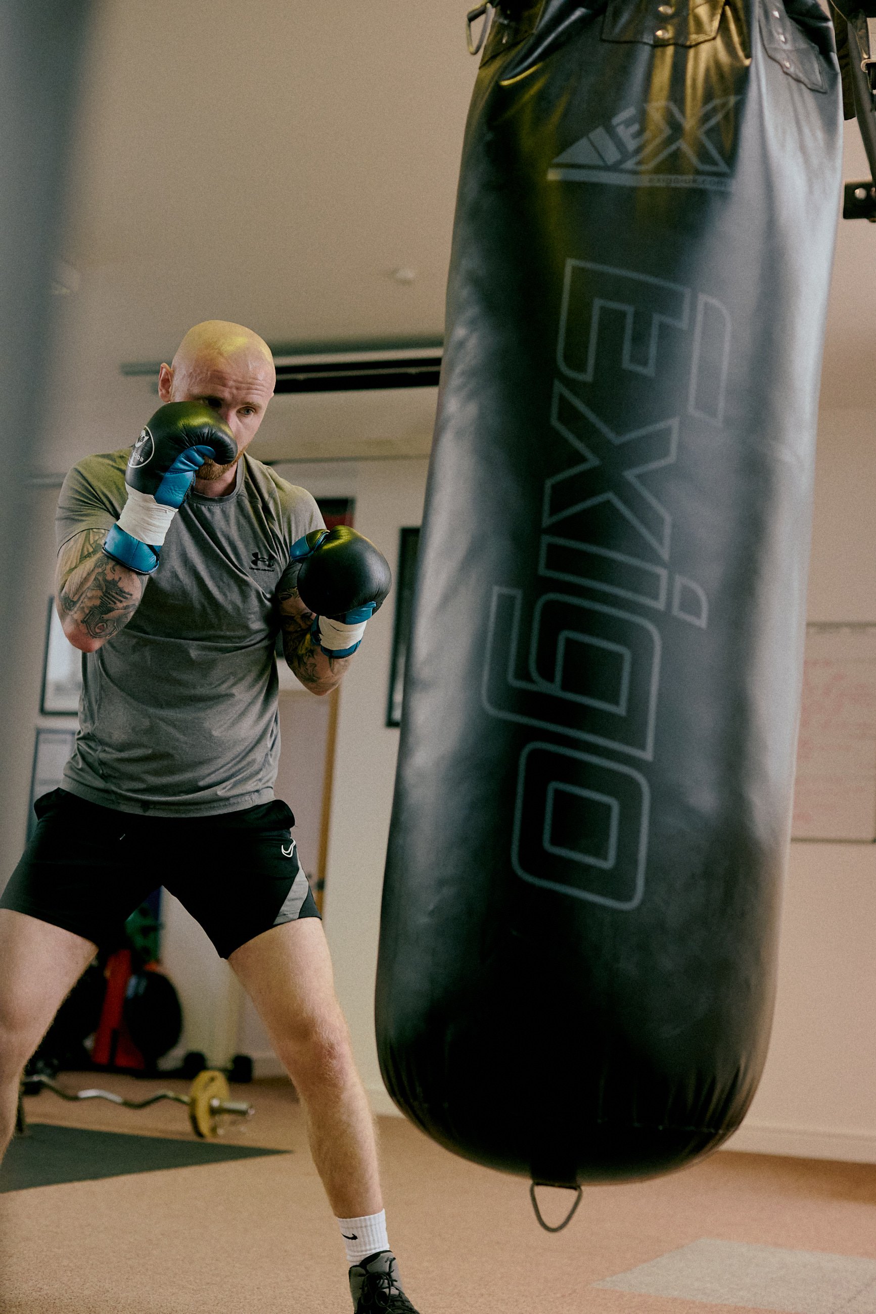 A man with a bald head, tattoos, wearing a gray athletic shirt, black shorts, and boxing gloves, is practicing boxing with a heavy punching bag labeled "BOXGLV" in a gym.