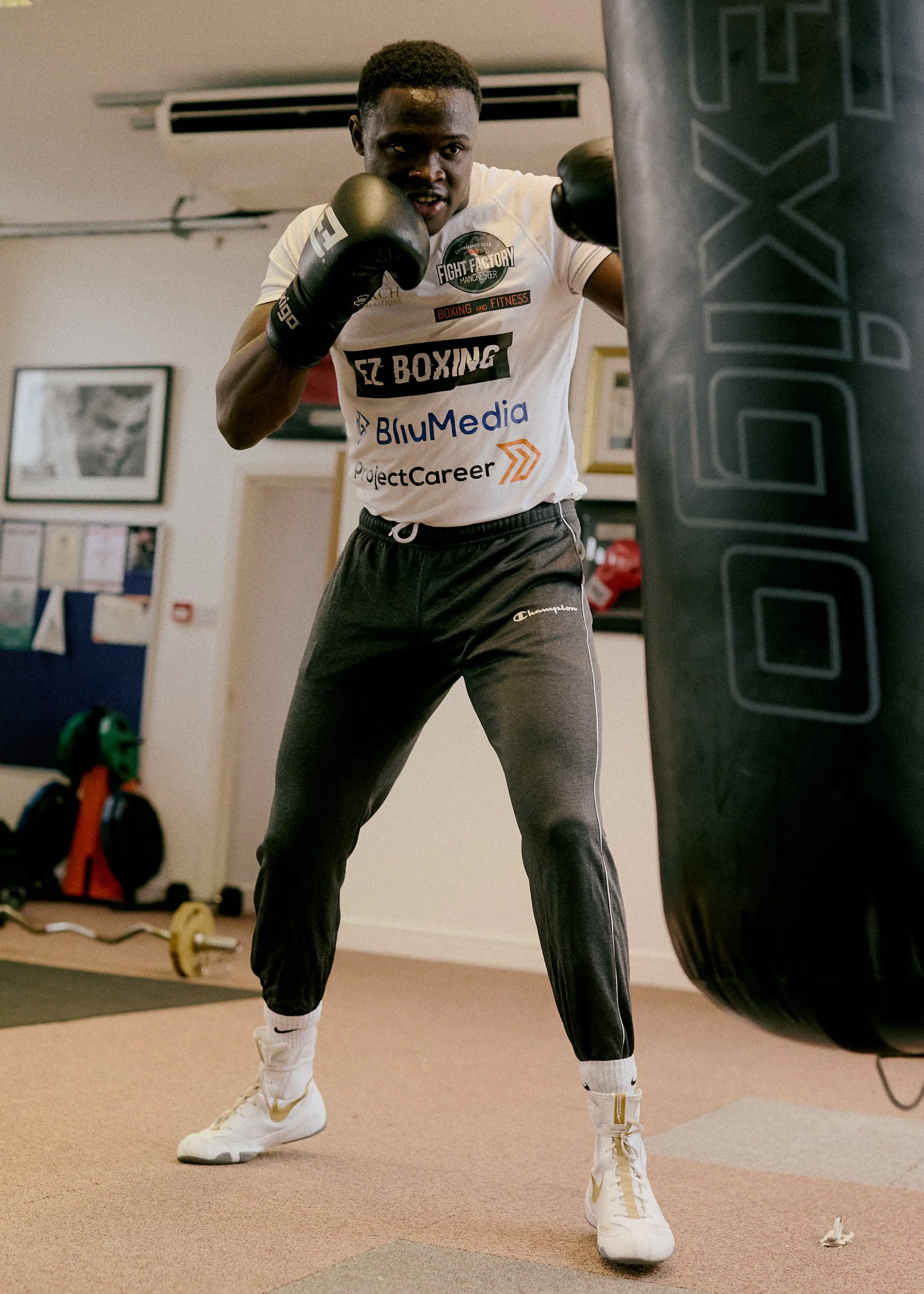 A young man throws a punch at a boxing heavy bag in a training room. He is wearing boxing gloves, a white t-shirt with various logos, black joggers, and white sneakers. The room has framed pictures on the wall and some gym equipment on the floor.