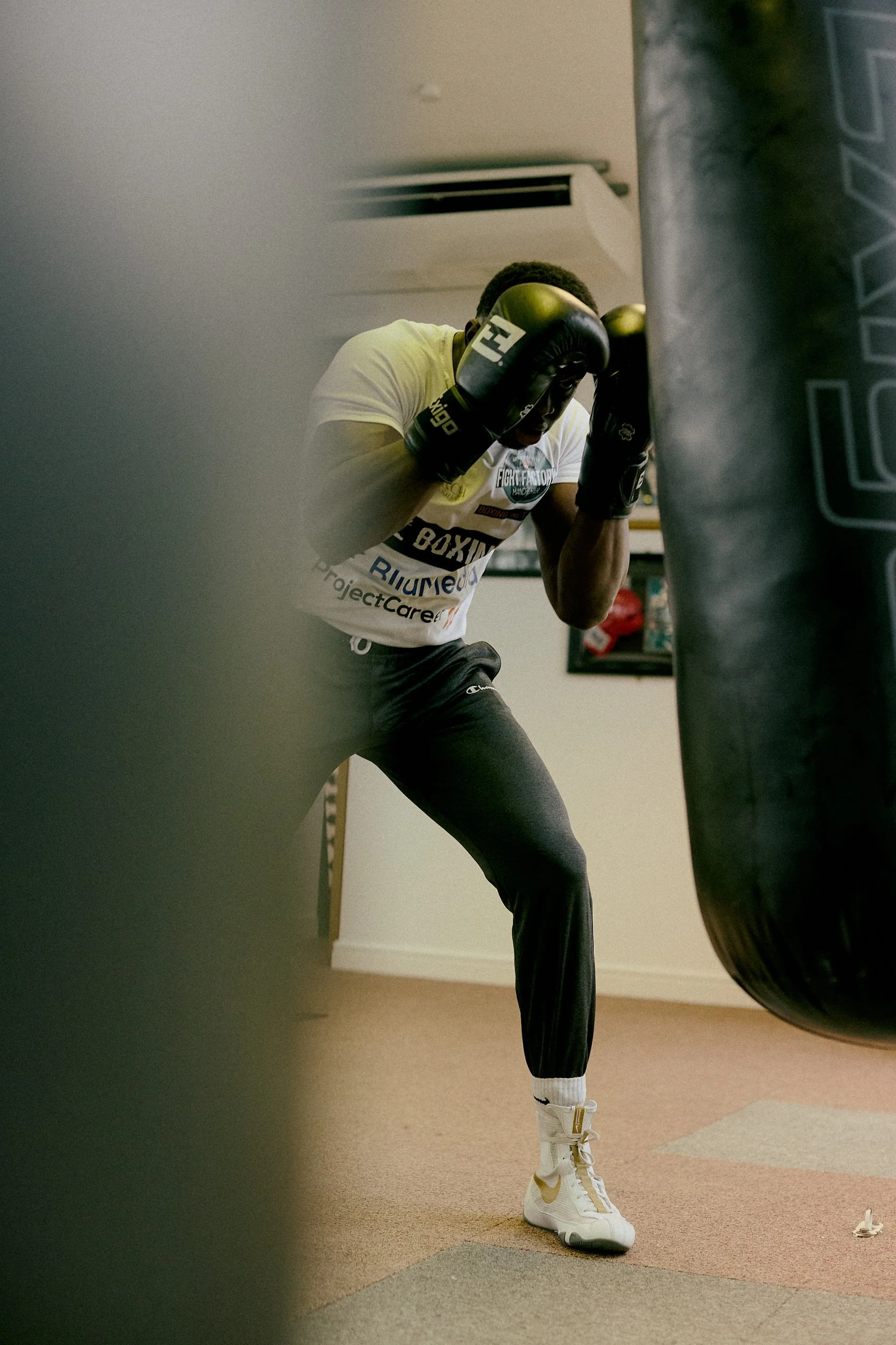 A young male boxer wearing boxing gloves, a T-shirt, and athletic pants practicing punching on a heavy bag in a gym.