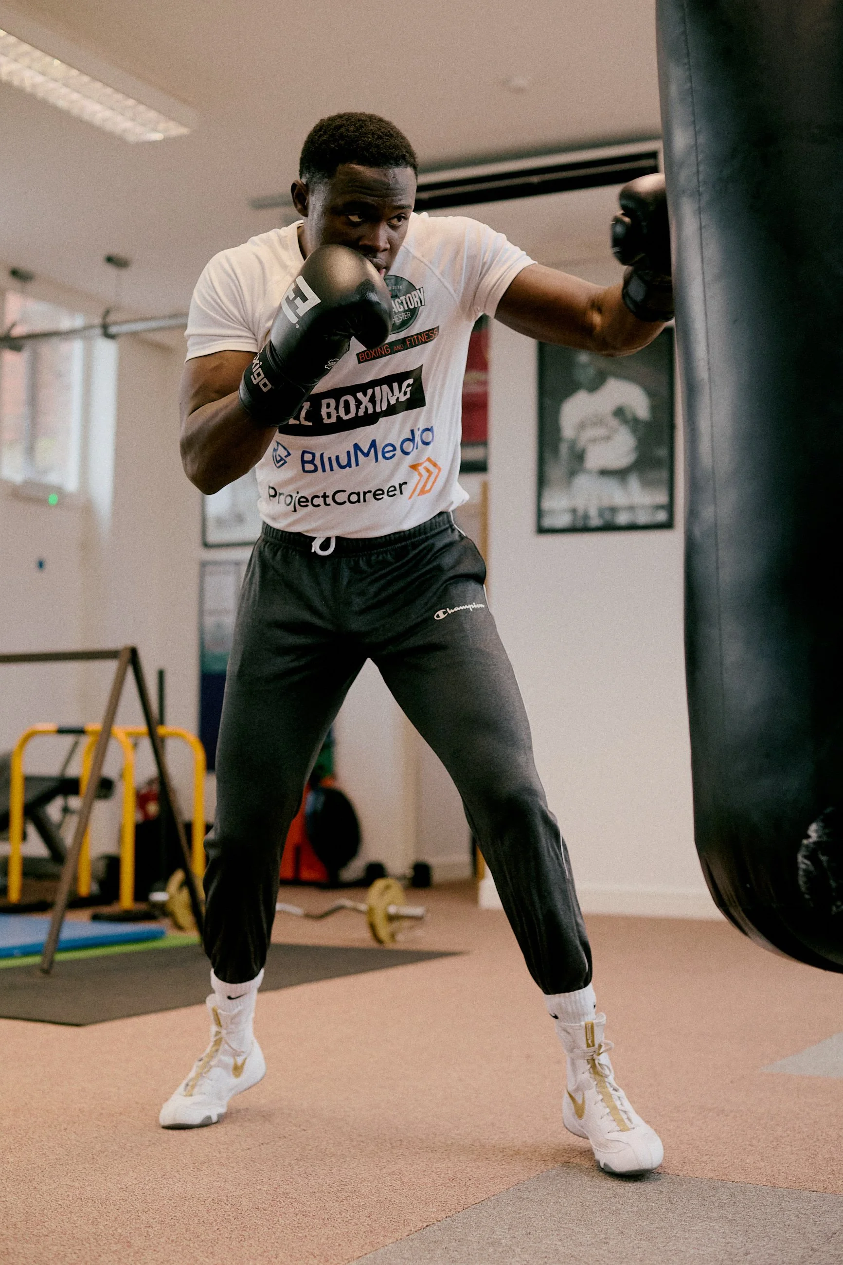 A young man wearing white Nike sneakers, black Champion sweatpants, and a white t-shirt with various logos, is practicing boxing in a gym. He is wearing black boxing gloves and is punching a black punching bag.