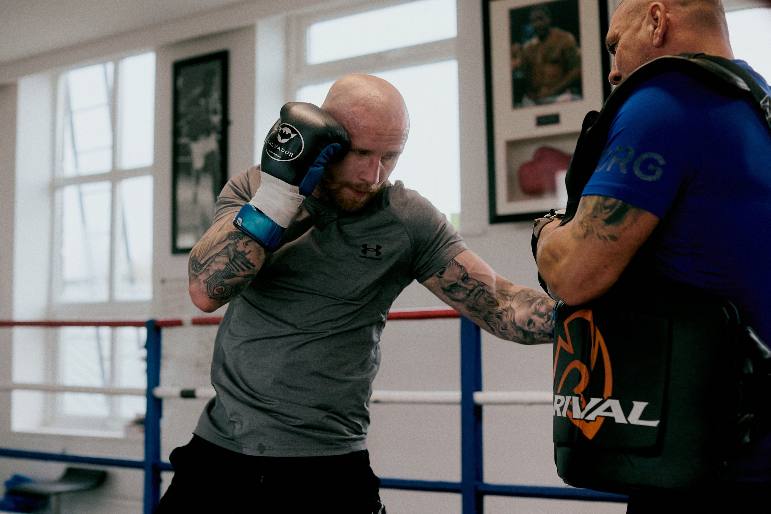 A boxer in a gray Under Armour shirt and boxing gloves practicing punches with a trainer in a gym.