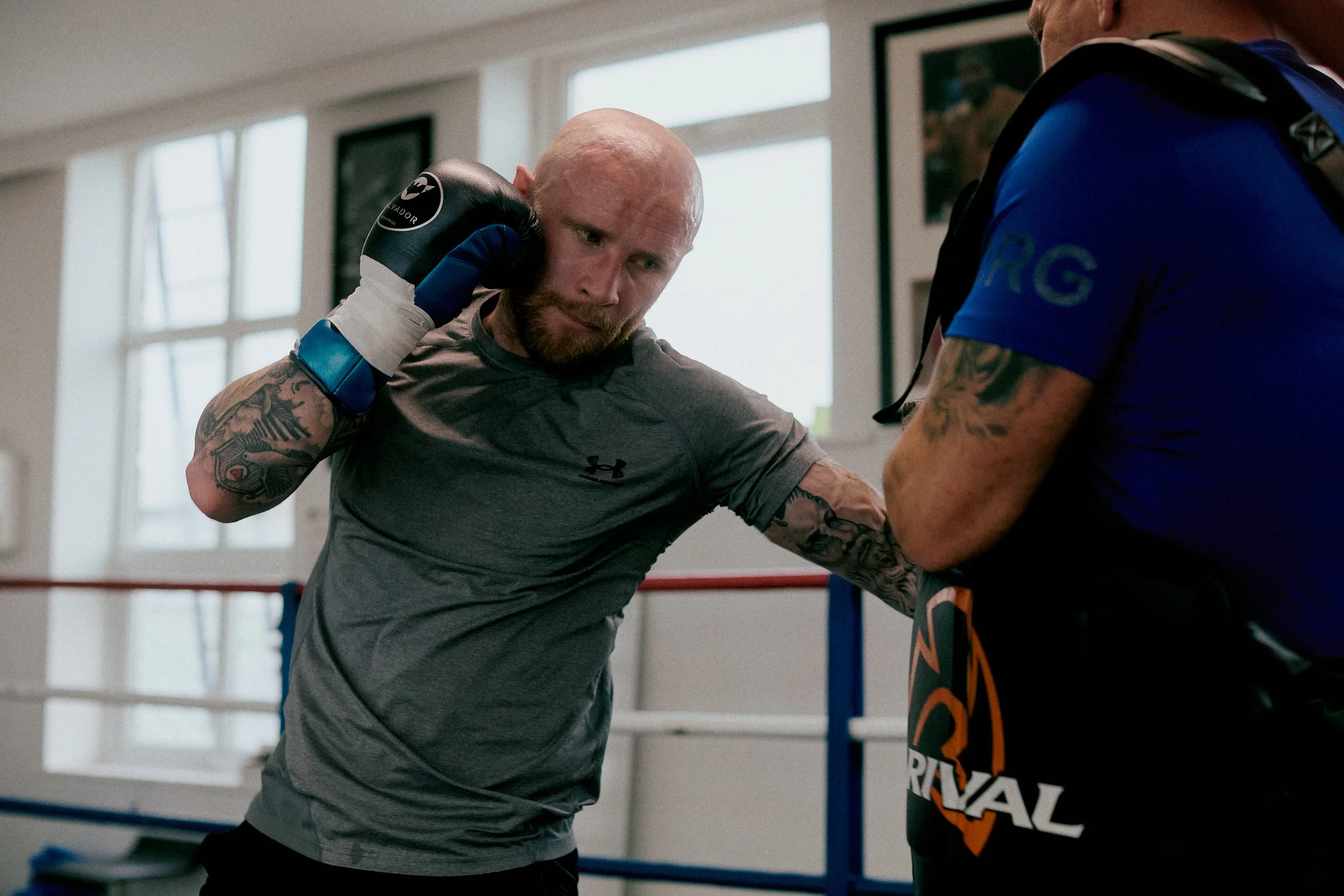A boxer in a gray shirt with tattoos on his arm punching a pad held by his trainer in a boxing gym.