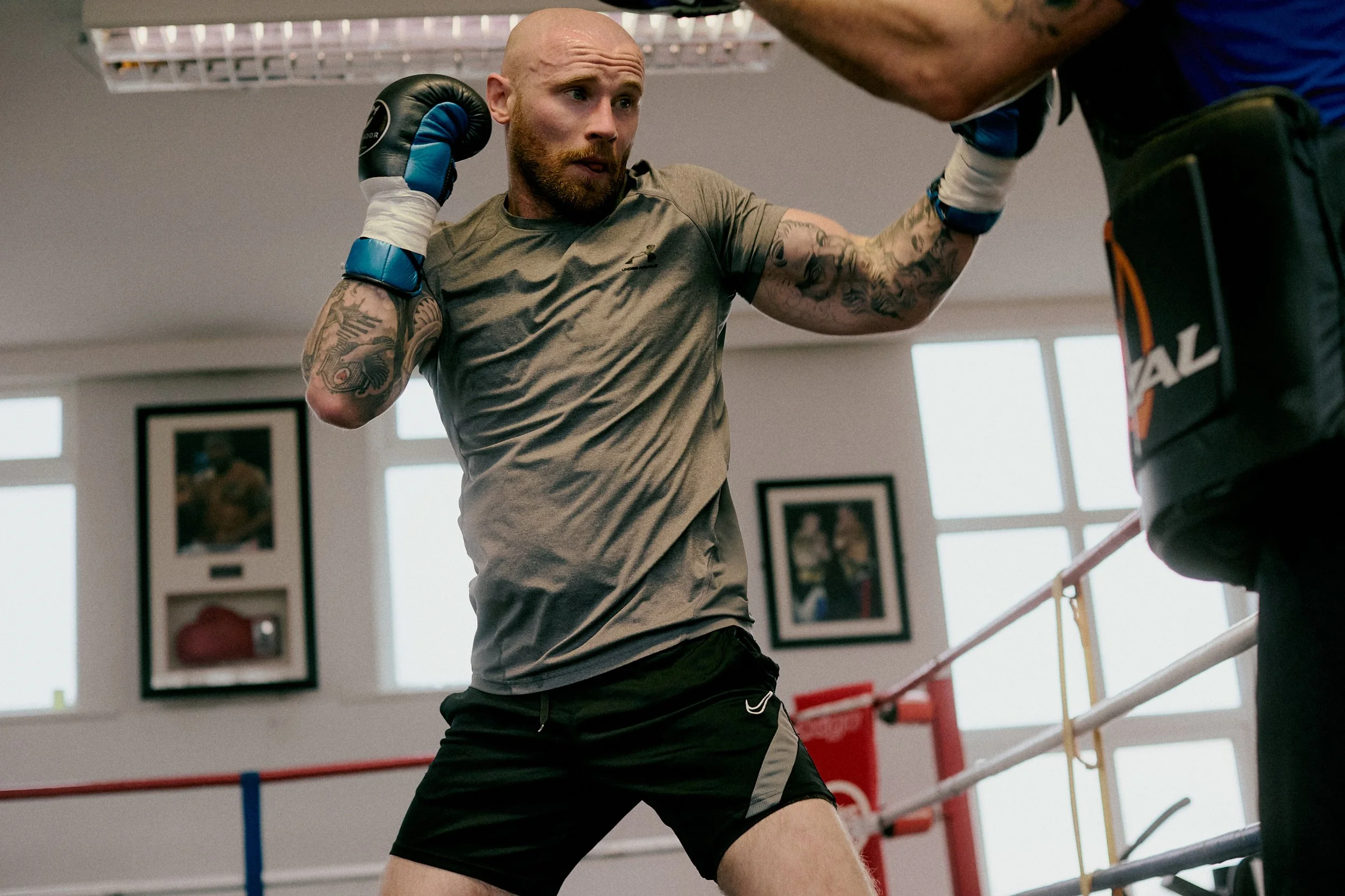 A man with tattoos in a gym, wearing a grey t-shirt and black shorts, practicing boxing with a trainer.
