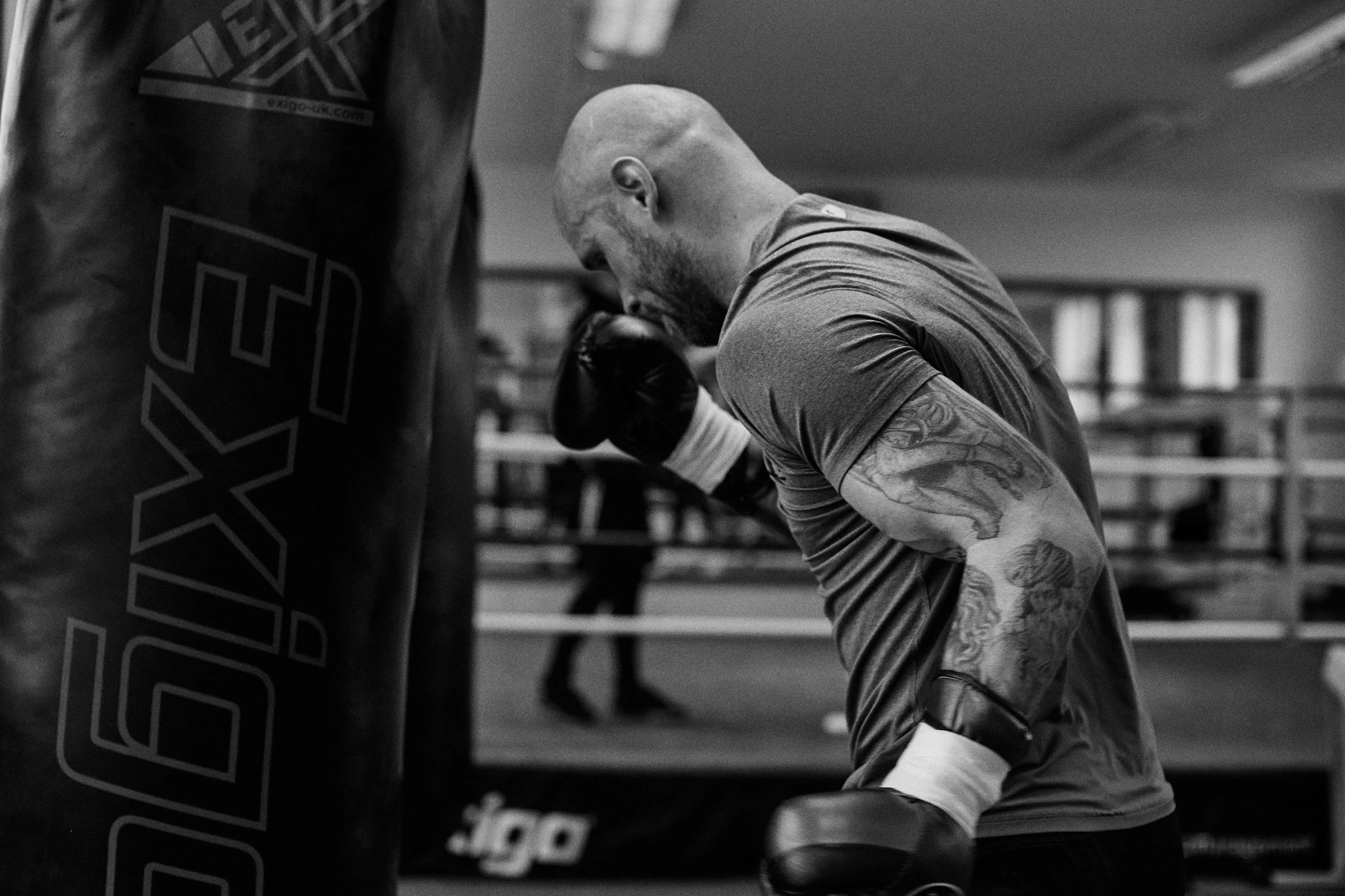 A male boxer with tattoos on his arms training in a gym, wearing gloves and throwing a punch towards a punching bag.