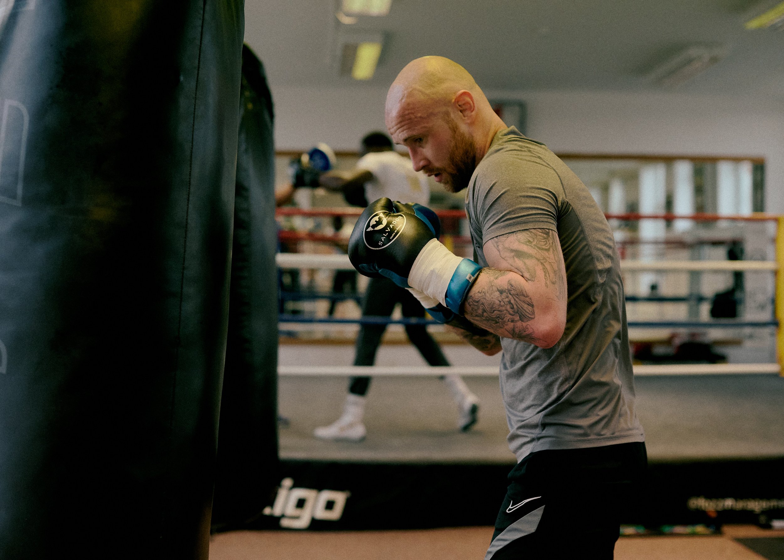 A man with tattoos on his arm practicing boxing in a gym, wearing gloves and positioned near a punching bag.