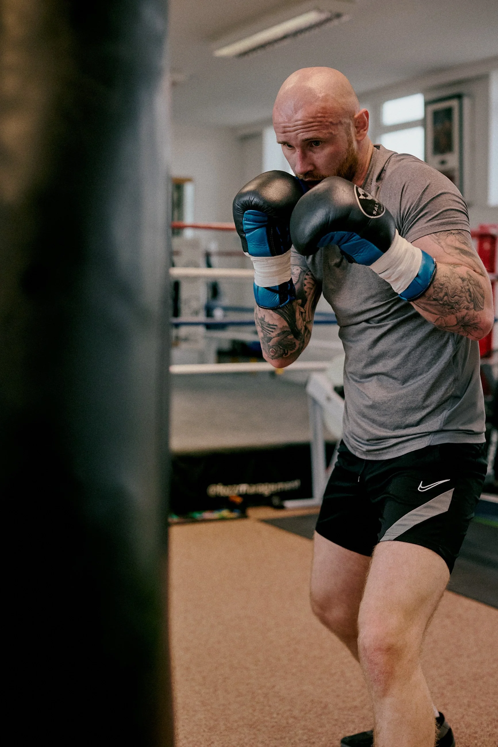 A man with a shaved head and tattoos on his arms wearing boxing gloves, a gray t-shirt, and black shorts practicing boxing in a gym, focusing on a punching bag.