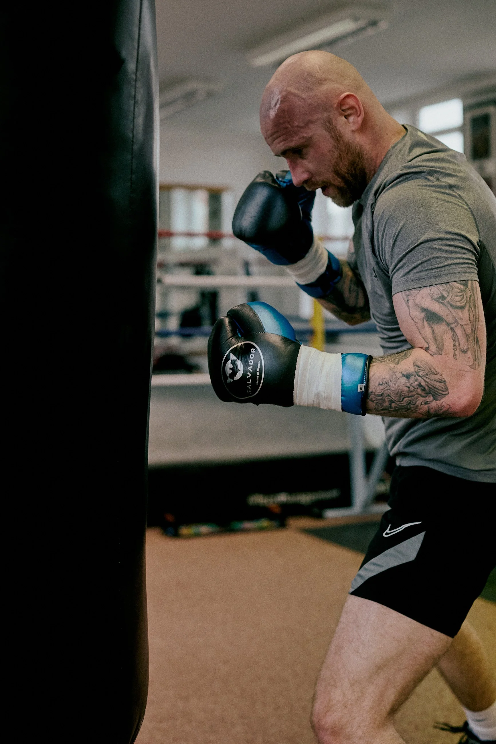 A man with tattoos practicing boxing on a punching bag in a gym, wearing boxing gloves and yoga shorts.
