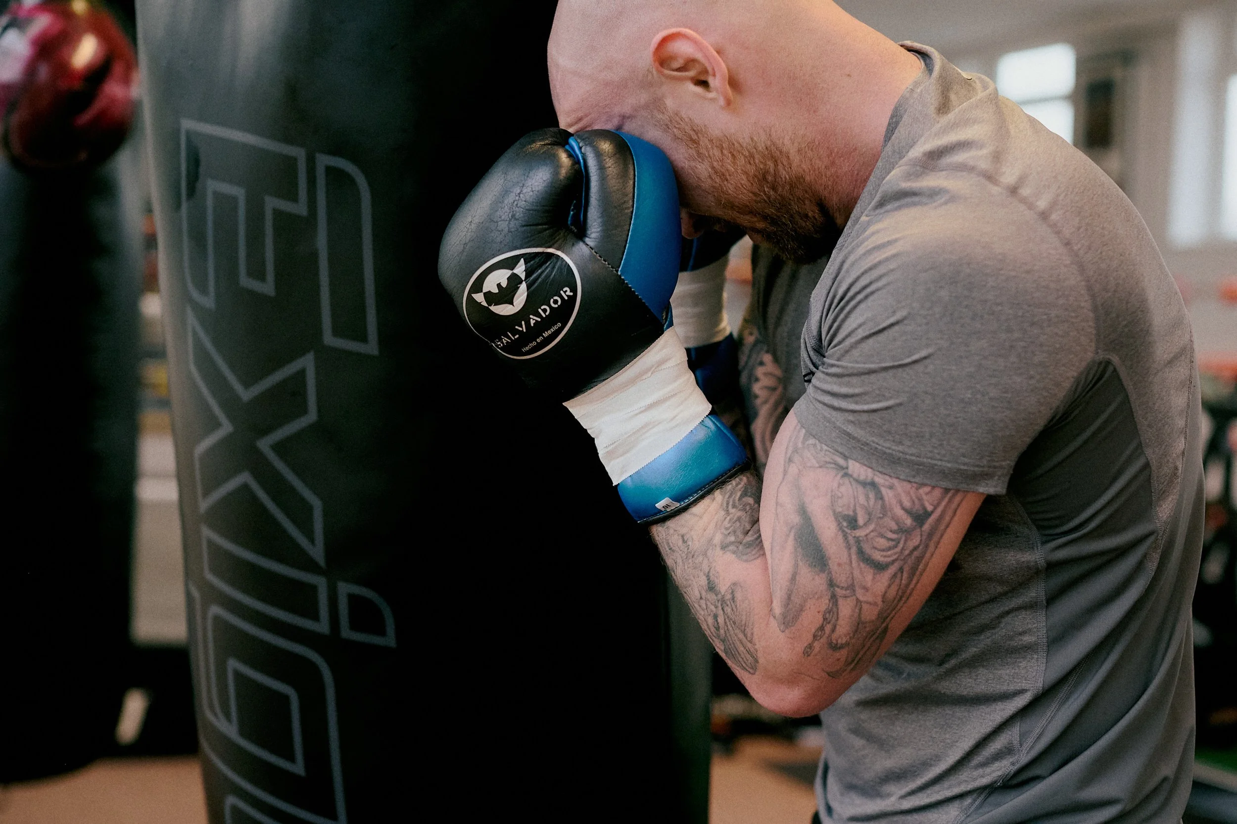 A male boxer with tattoos on his left arm rests his head and forehead against a black punching bag in a gym, wearing boxing gloves and a gray athletic shirt.