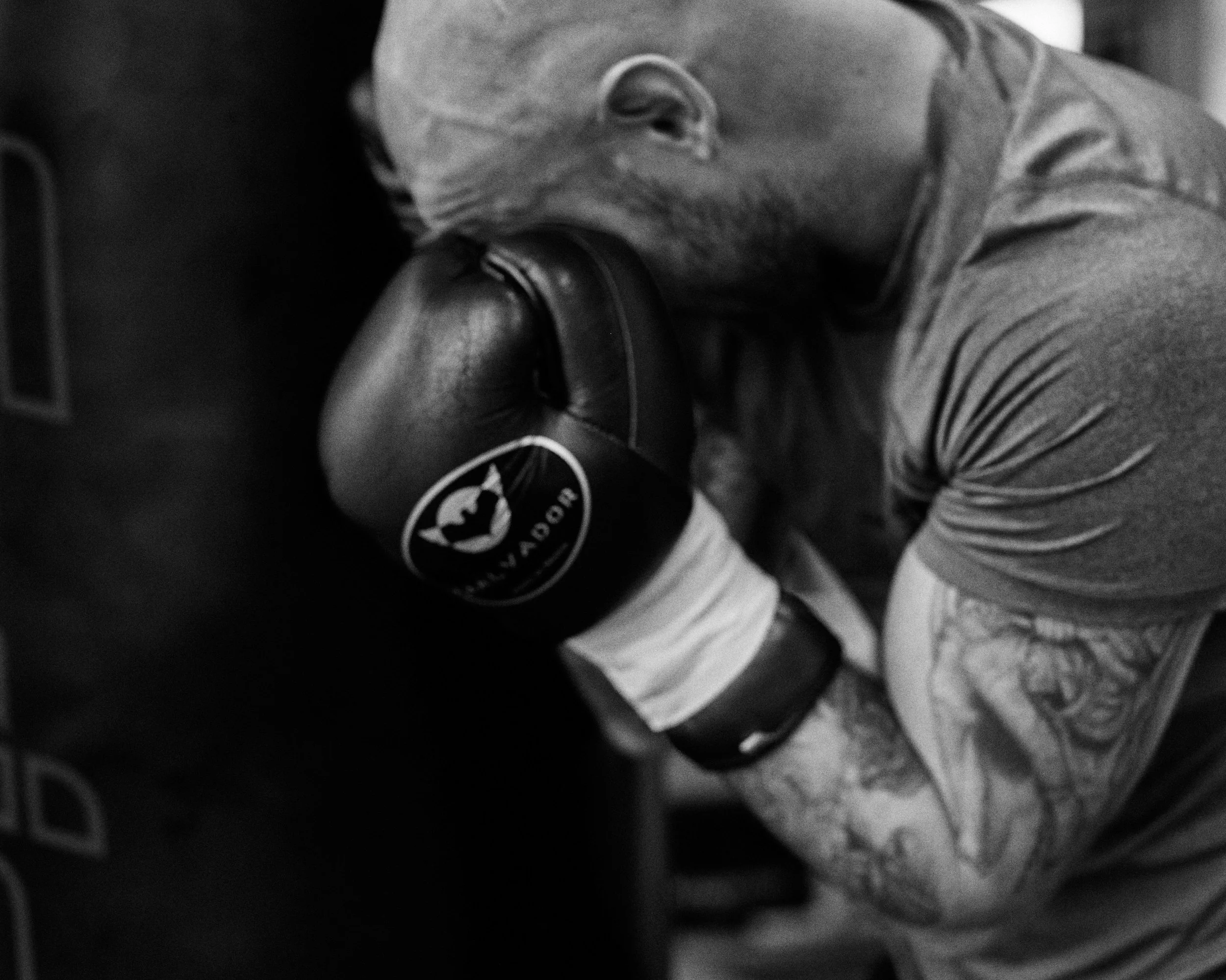 A man with tattoos on his arm and a shaved head is resting his face on a boxing glove, wearing a focused or tired expression.