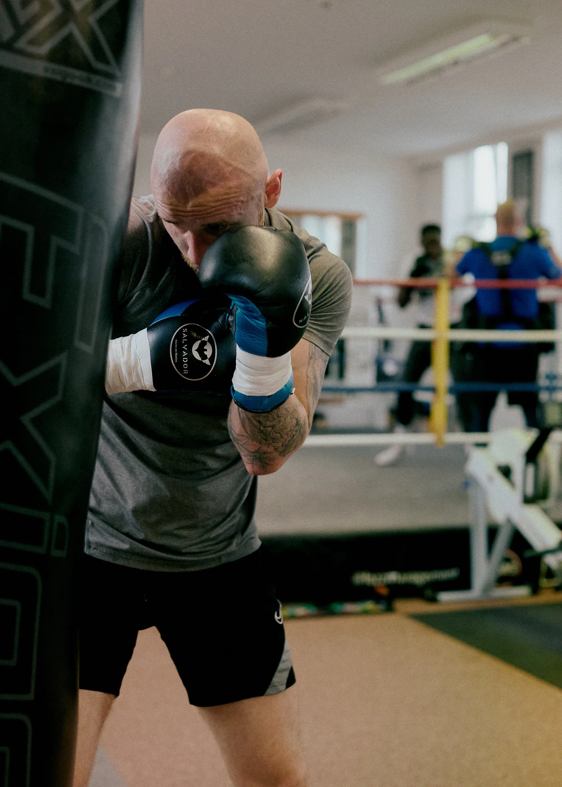 A man with a shaved head practicing boxing in a gym, leaning against a punching bag with his fists up.