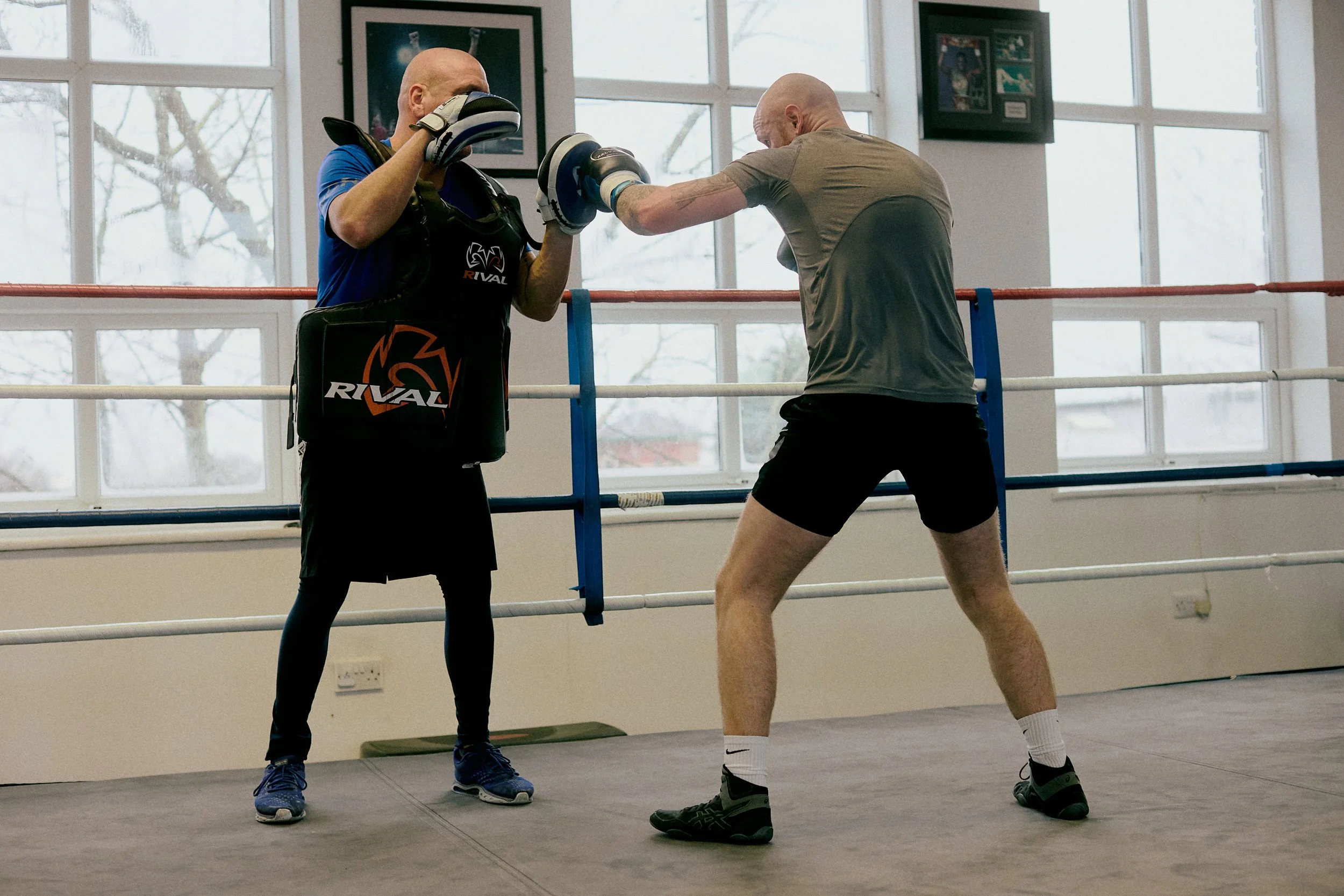 Two men practice boxing in a gym. One man on the left holds focus mitts, and the other man on the right is throwing a punch. The scene takes place inside a boxing ring with large windows in the background.