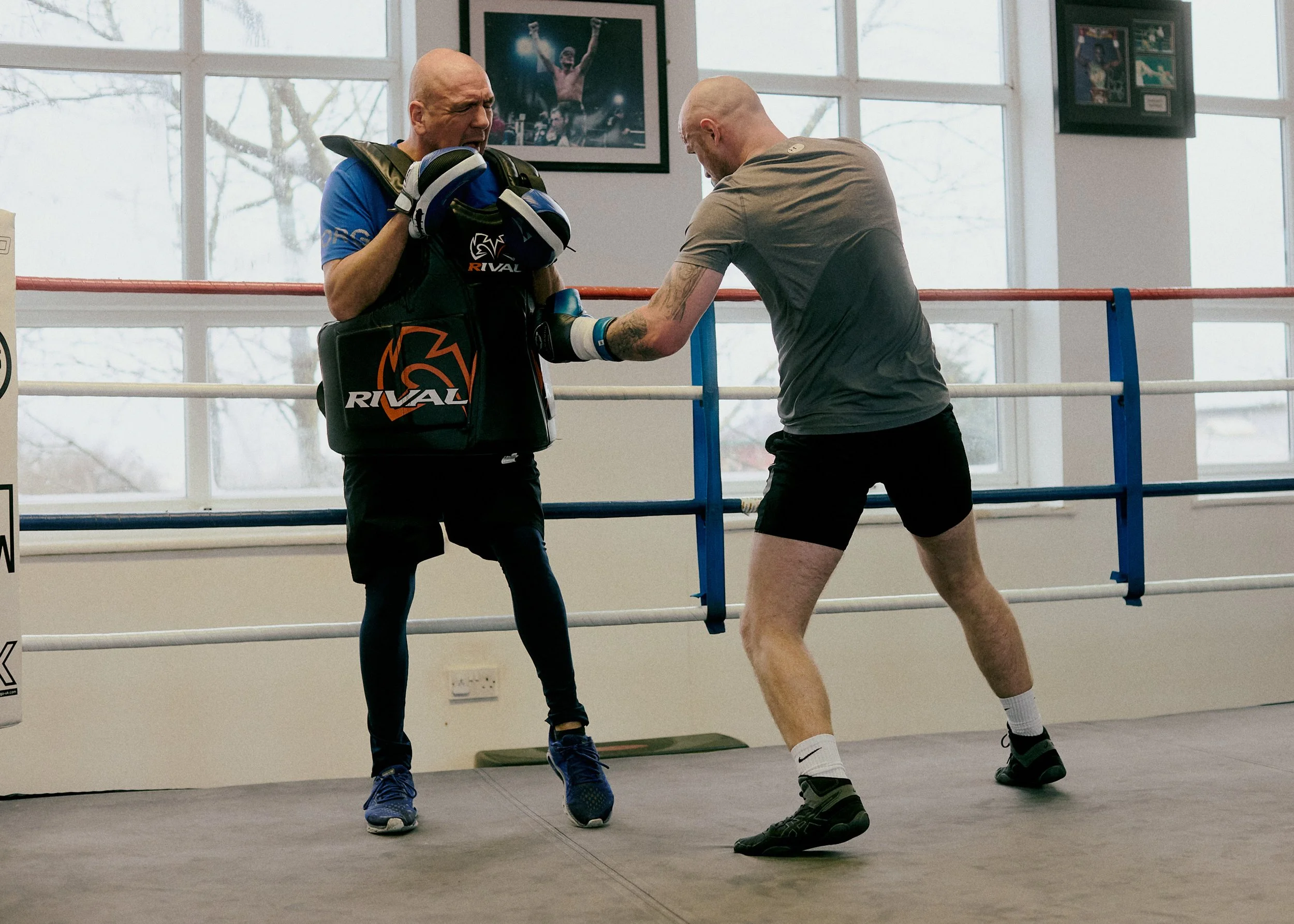 Two men training in a boxing gym, one with boxing gloves and pads, the other practicing punches, inside a boxing ring with framed photos on the wall.