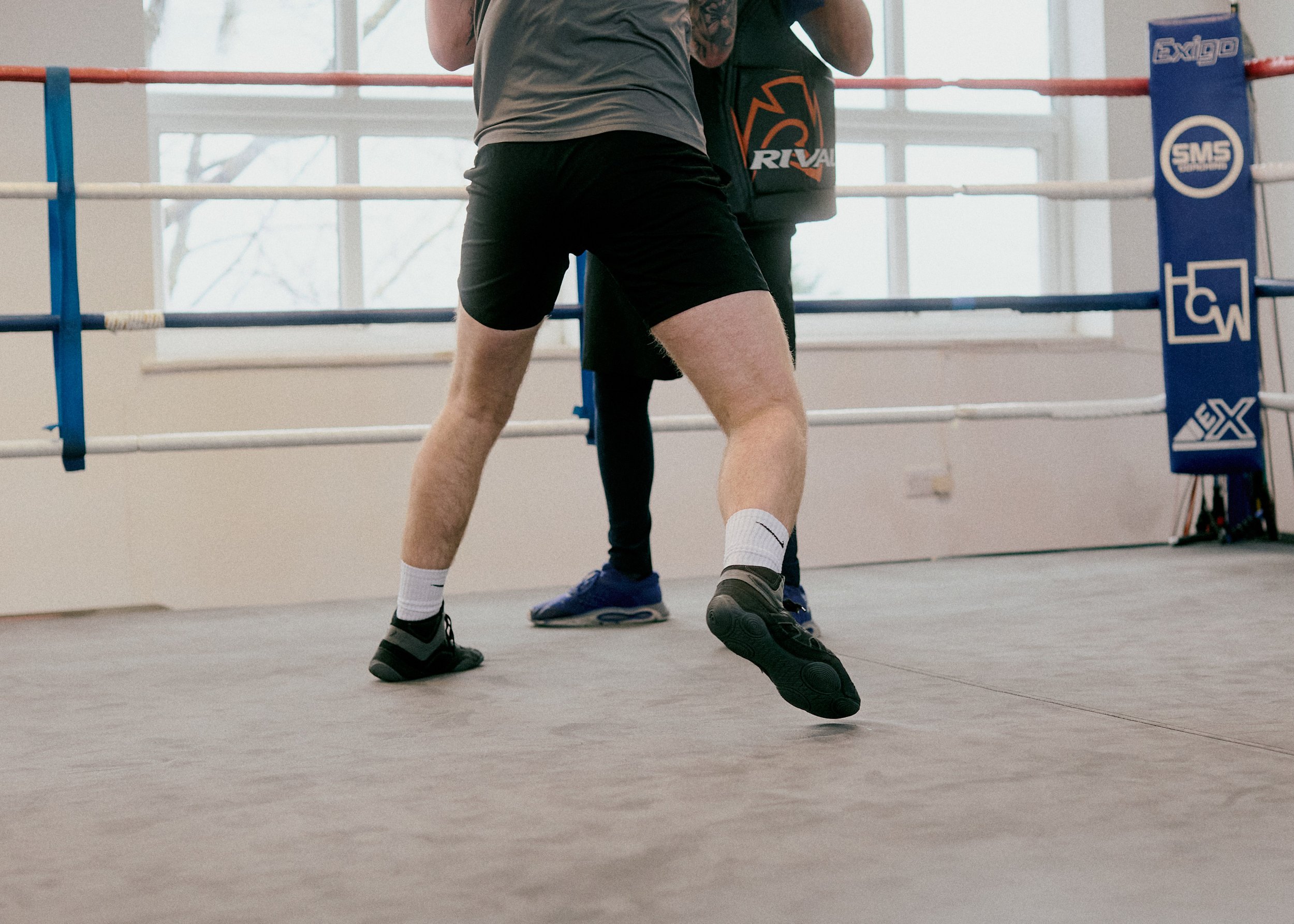Two people in a boxing ring practicing boxing. One person is in a boxing stance with their legs apart, wearing black shorts, a gray t-shirt, and black shoes with white socks. The other person is standing behind them, wearing black pants and a black s