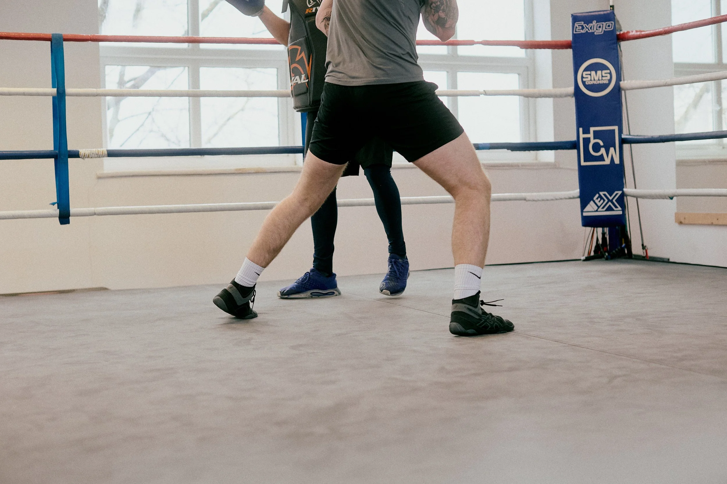 Two people boxing in a gym, standing in a fighting stance inside a boxing ring with ropes and padded corner posts.