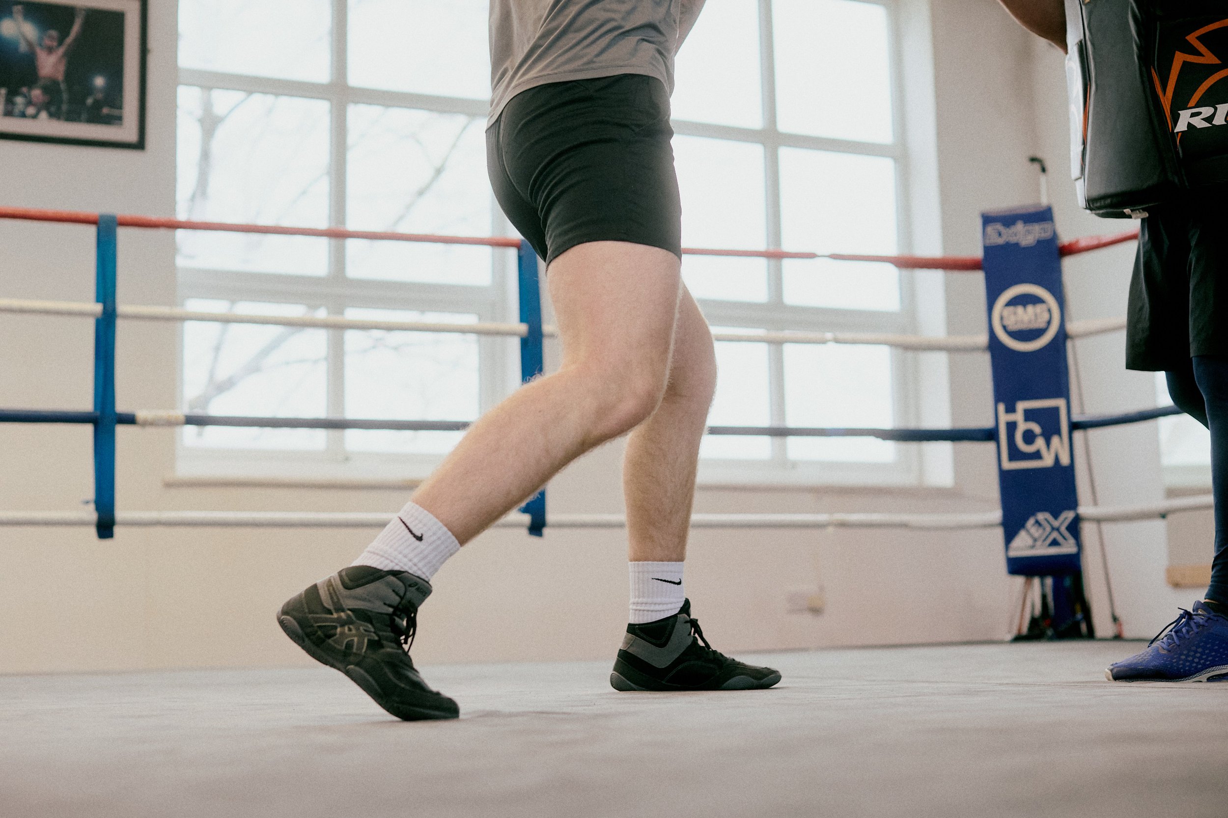 Close-up of a boxer practicing in a gym, showing his legs and part of his torso, with a boxing ring and trainer nearby.