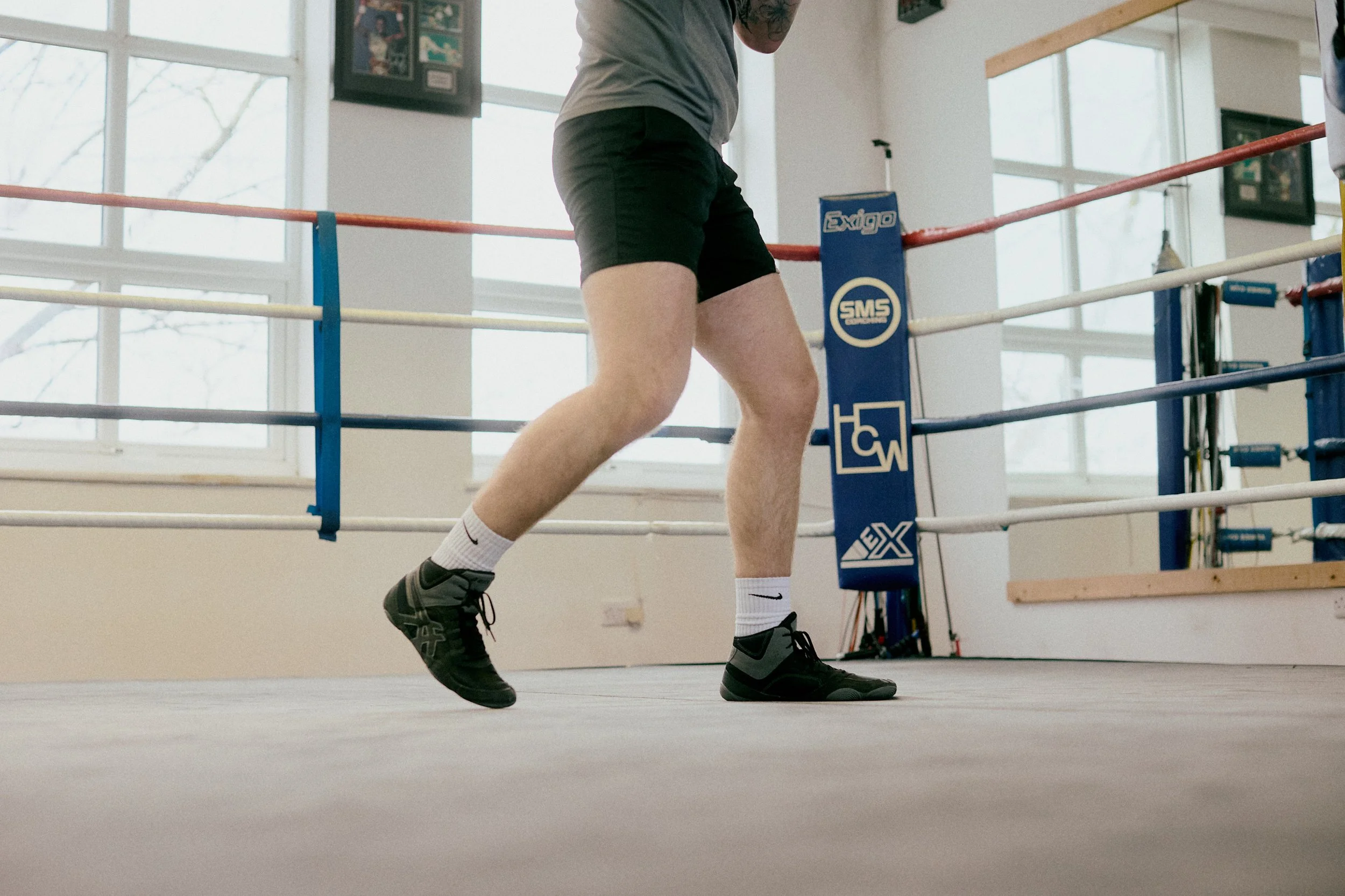 A person in athletic shorts and sneakers practicing boxing in a gym with a boxing ring, large windows, and fitness equipment.