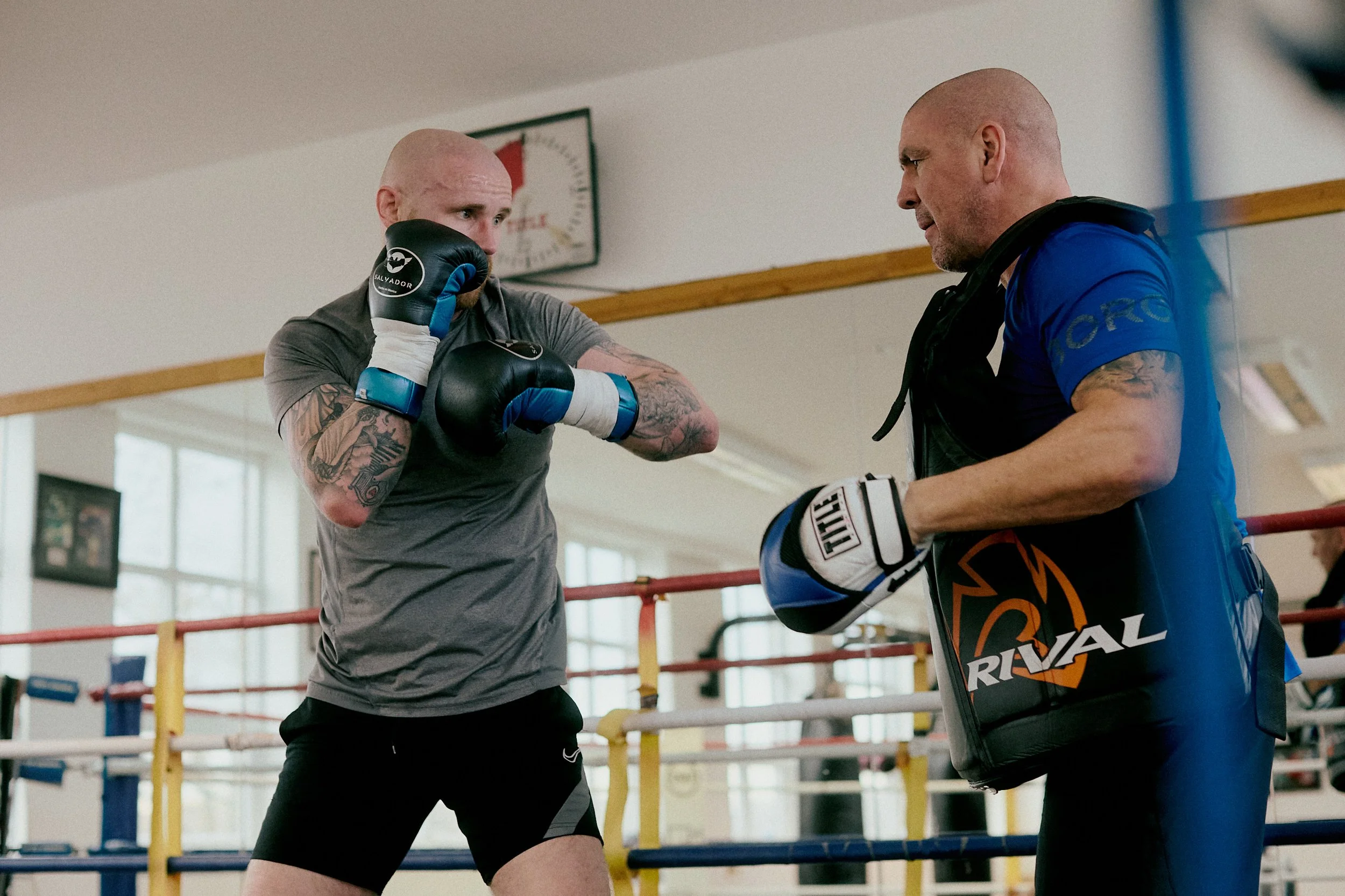 A boxer with tattoos stands in a boxing ring, wearing gloves and gray shirt, practicing with a trainer who is holding focus mitts in a gym.