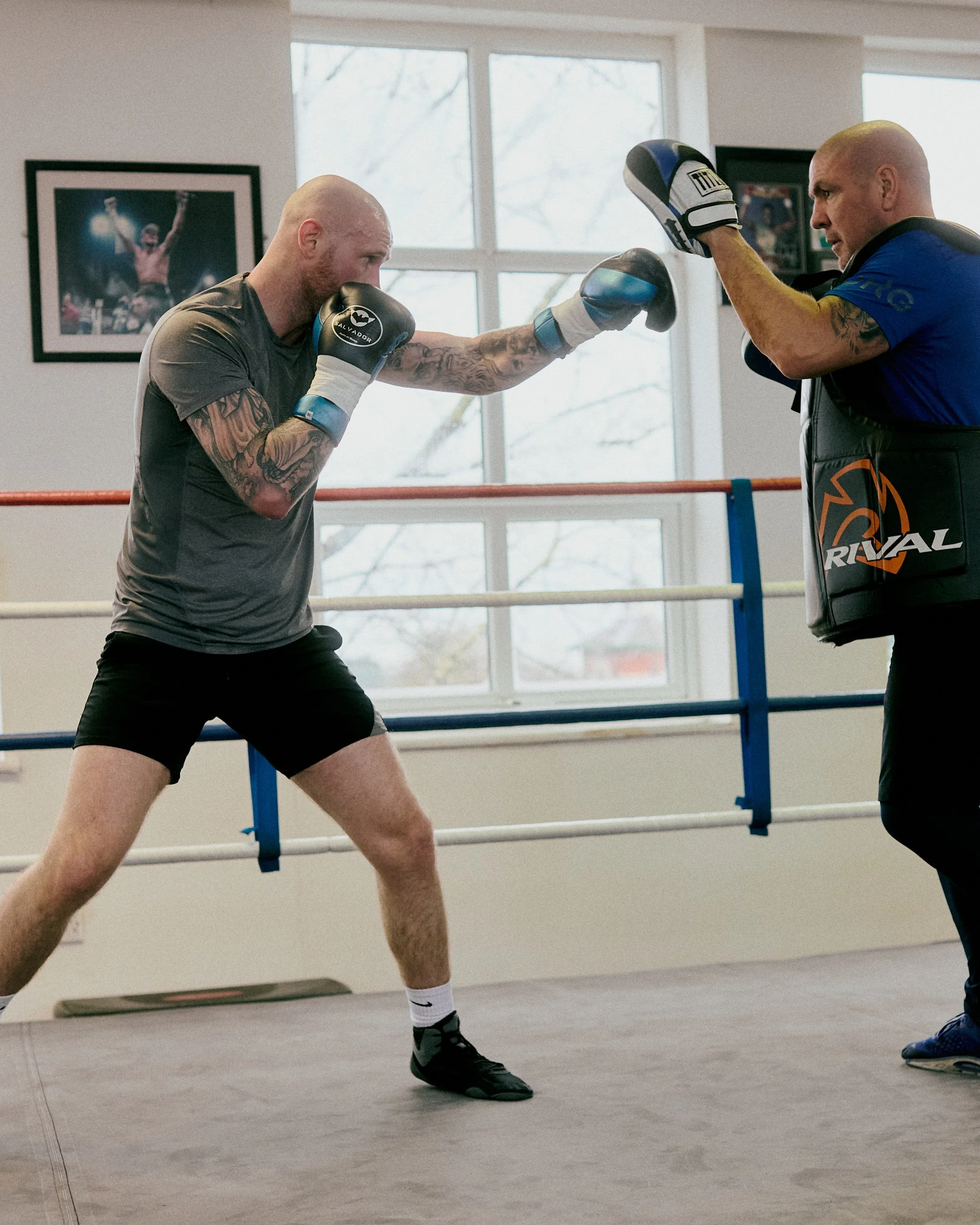 A boxer in gray shirt and black shorts practicing boxing with a punch mitt trainer wearing a blue shirt and holding focus mitts in a boxing gym.