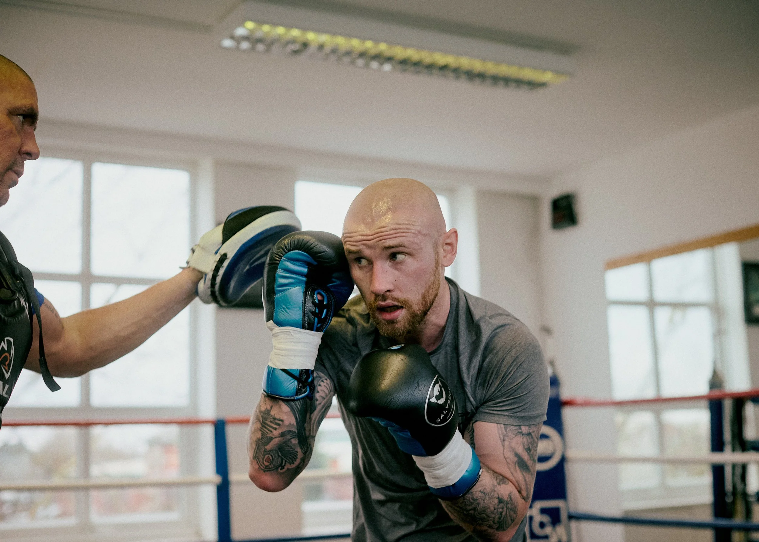 A boxer in boxing gloves practicing punches with a trainer in a boxing gym, with large windows in the background.