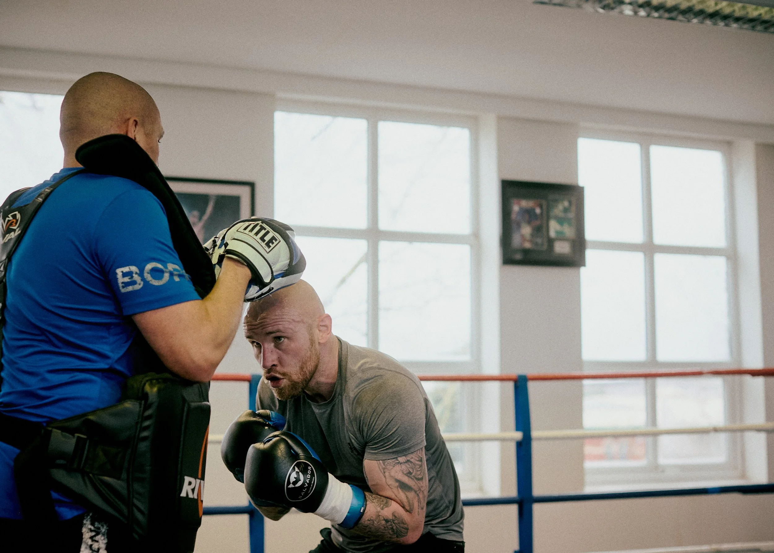 A bald man wearing boxing gloves and a gray t-shirt practices boxing with a trainer in a gym with large windows.