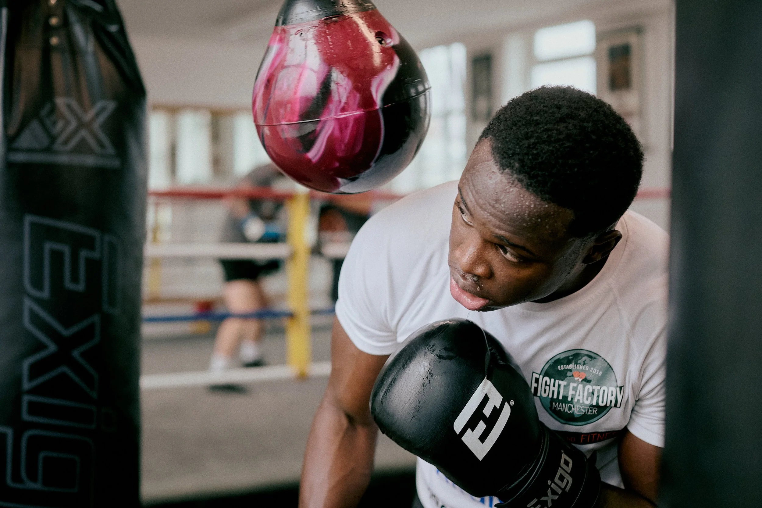 A young male boxer in a gym, wearing gloves and a t-shirt, practicing punches on a boxing bag.